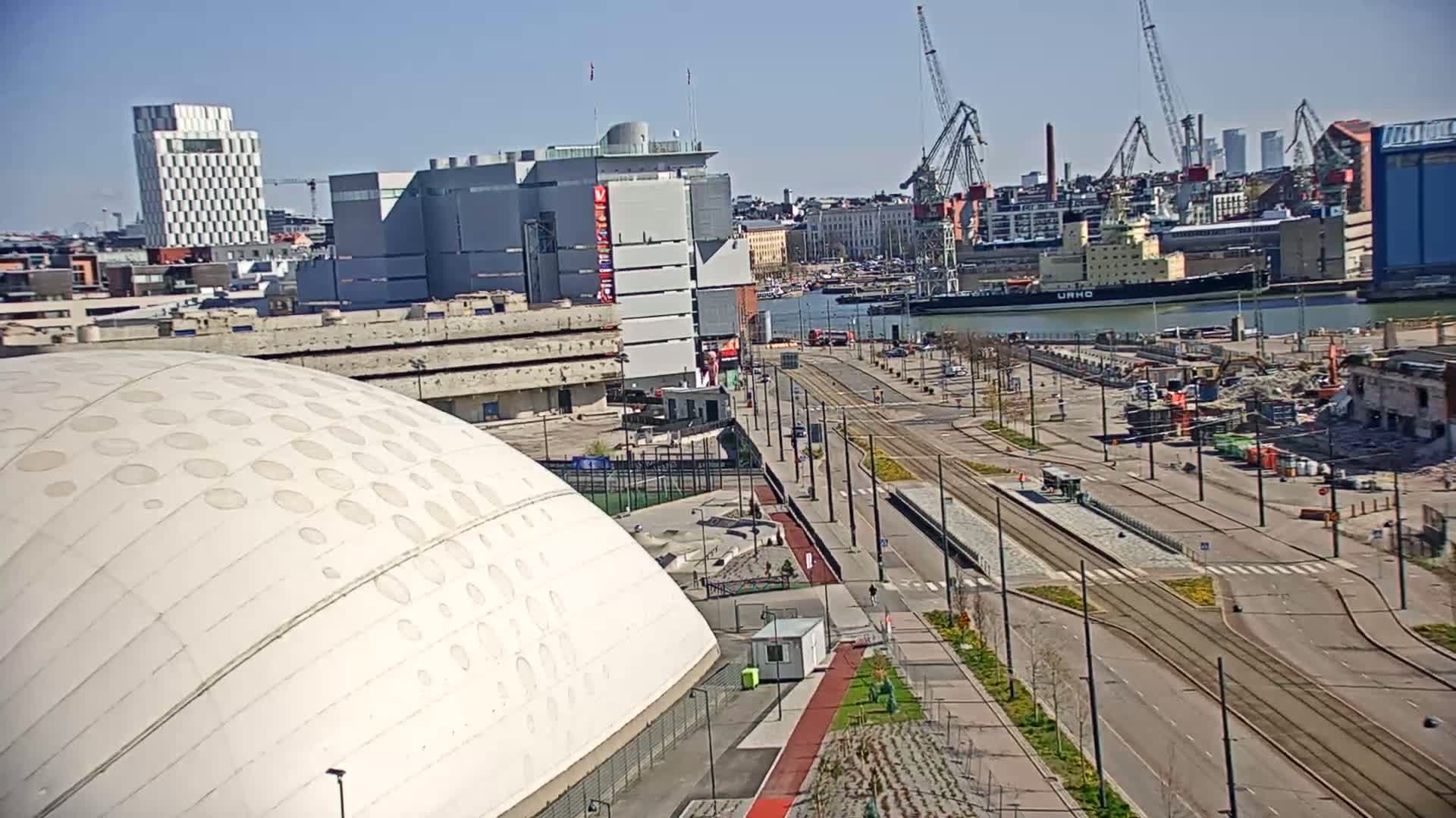 A high-angle view on a sunny day shows a large white domed building next to a city street with tram tracks, construction activity, and a harbor with several cranes and ships in the background.