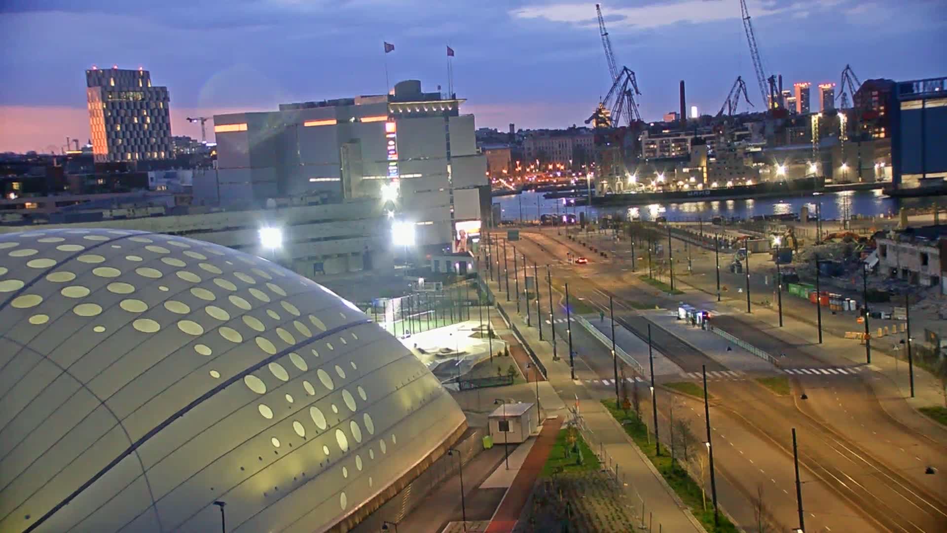 A large domed building with circular cutouts is situated near a waterfront city at twilight, with a wide, mostly empty road and harbor visible in the background.