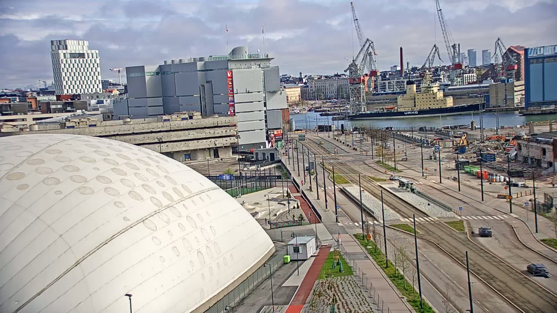 A partly cloudy day overlooking a cityscape with a large white domed building in the foreground, tram tracks and roadways leading to a harbor with a large ship and construction cranes.