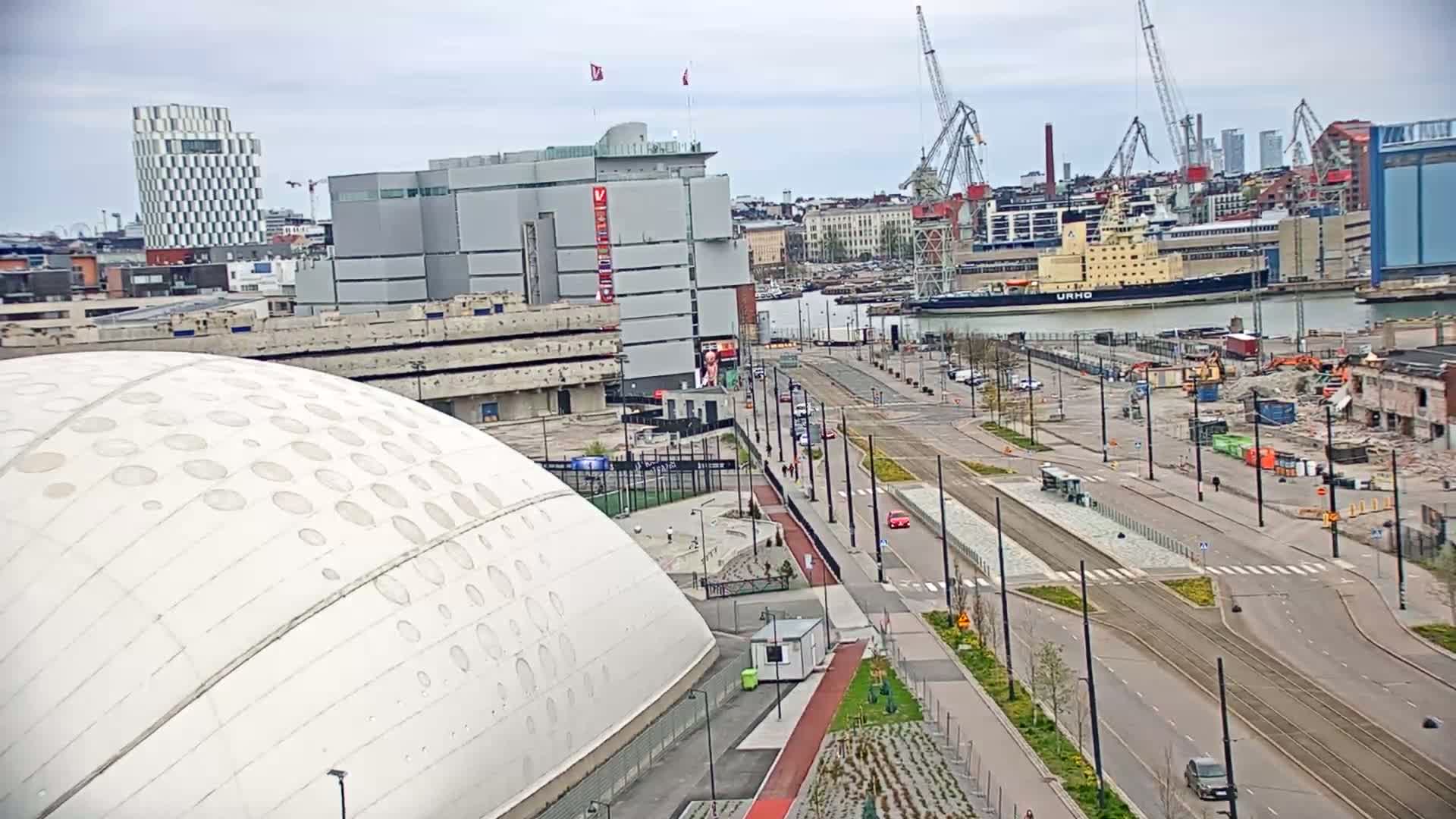 A large white domed building sits beside a wide, mostly empty street with tram tracks running down the center, overlooking a harbor with a large ship and construction cranes under a cloudy sky.