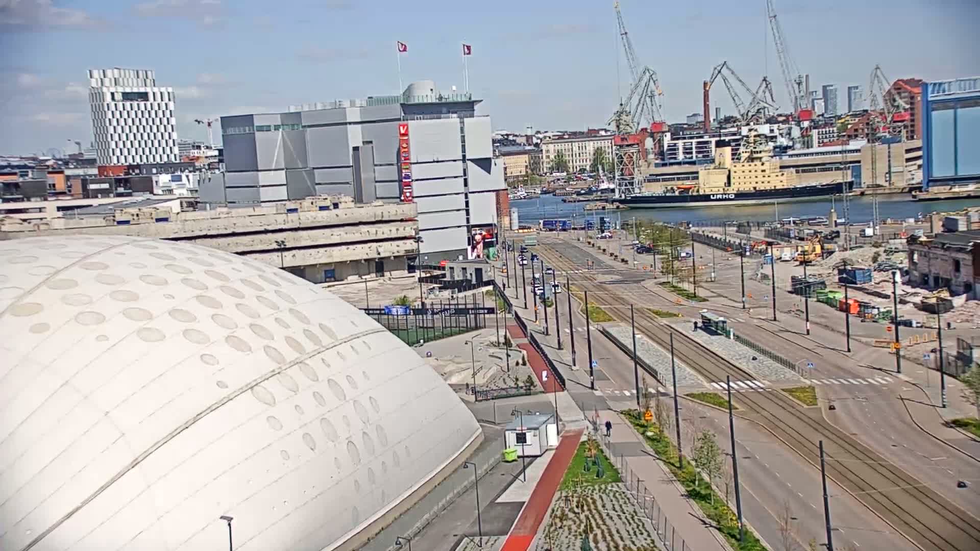 A high-angle view on a sunny day shows a large domed building next to a wide roadway with tram tracks, a harbor with ships, and a cityscape in the background.