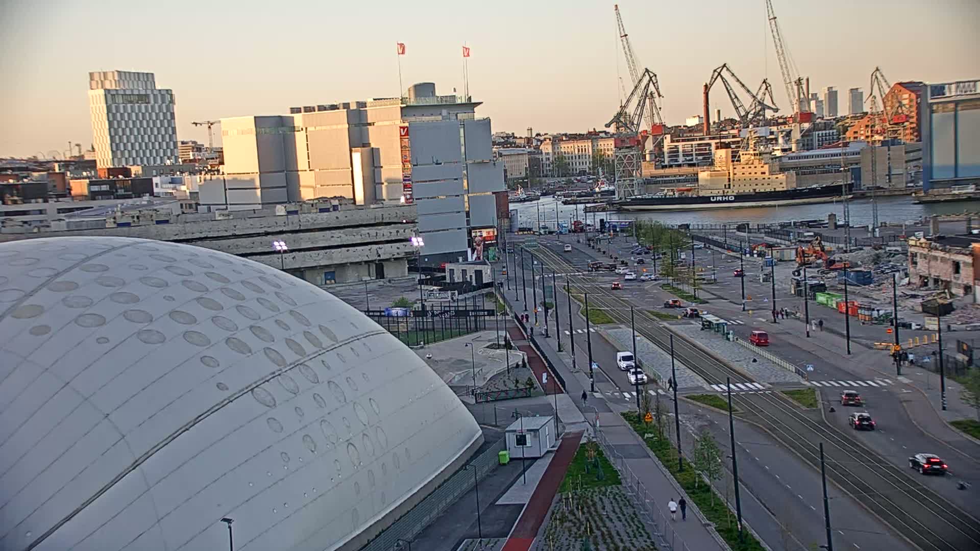A large domed building sits beside a city street with tram tracks, overlooking a harbor with several cranes and a large ship at sunset.