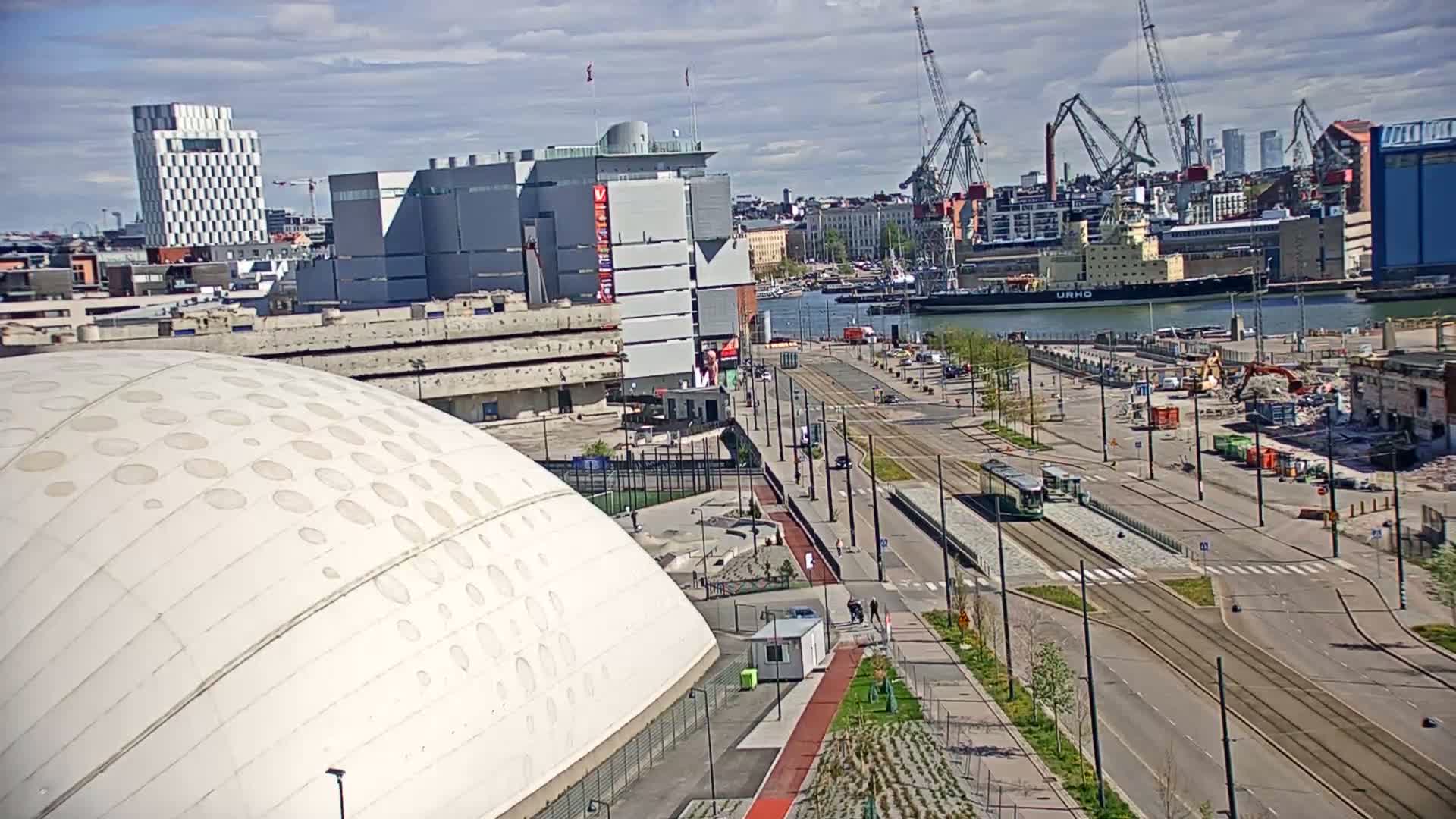 A large domed building is situated near a harbor on a partly sunny day, with a tram running along a road next to a construction site and a body of water visible in the background.