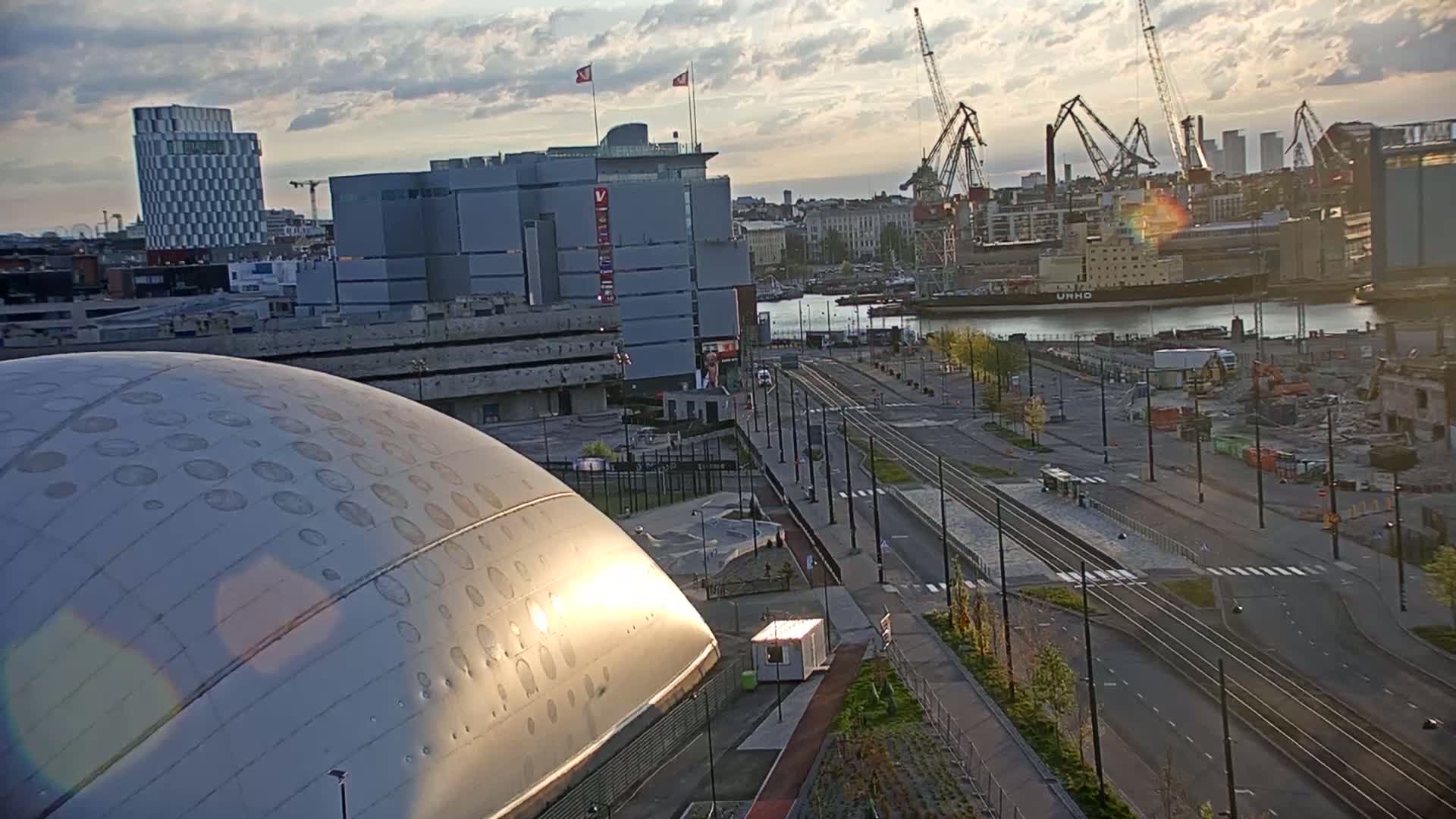 A large, domed building sits in the foreground, overlooking a city street with tram tracks, a harbor with ships and cranes, and other buildings under a partly cloudy sunset sky.
