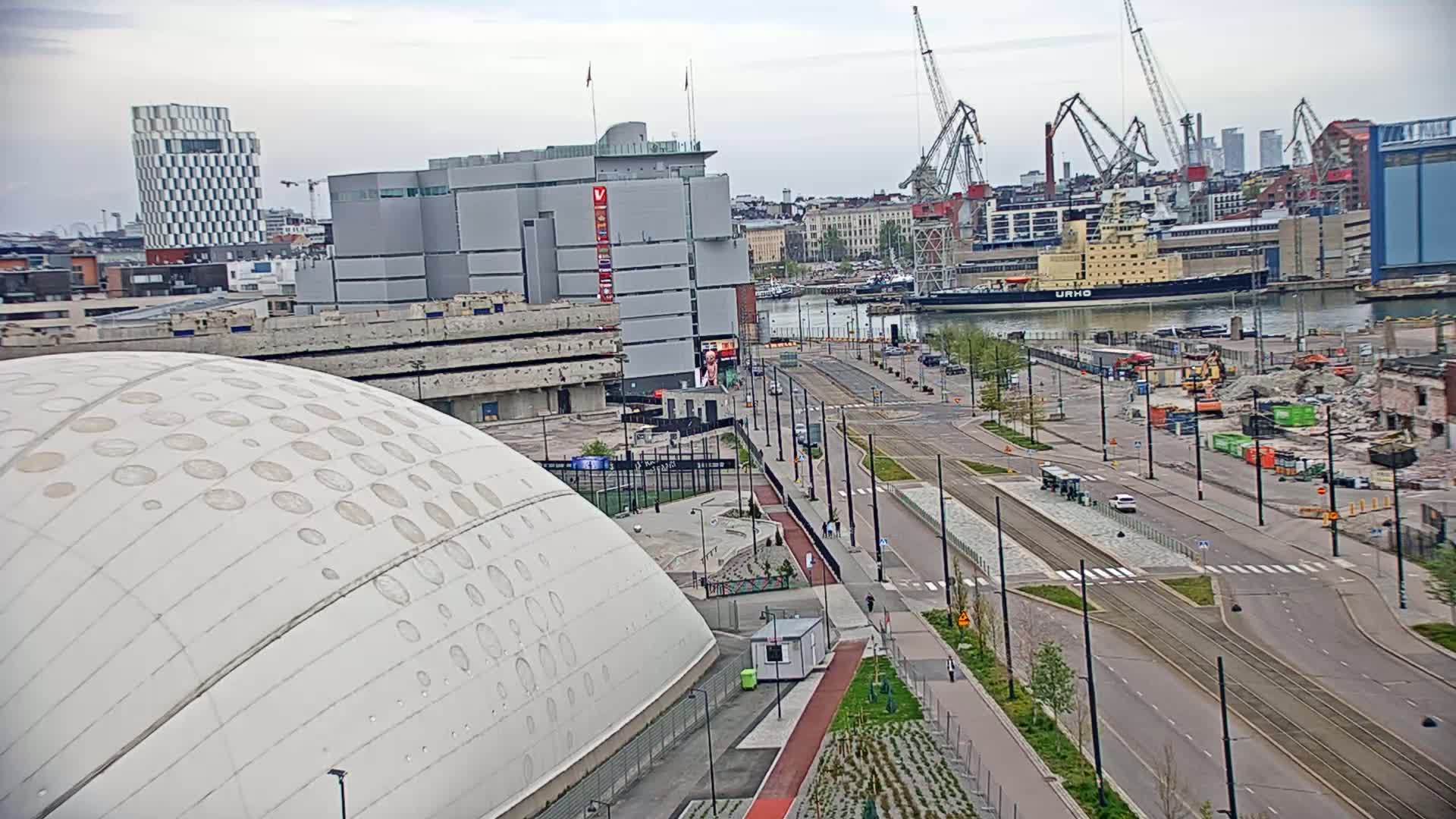 A large, domed building sits next to a wide city street with tram tracks, overlooking a harbor with a large ship and active cranes under an overcast sky.