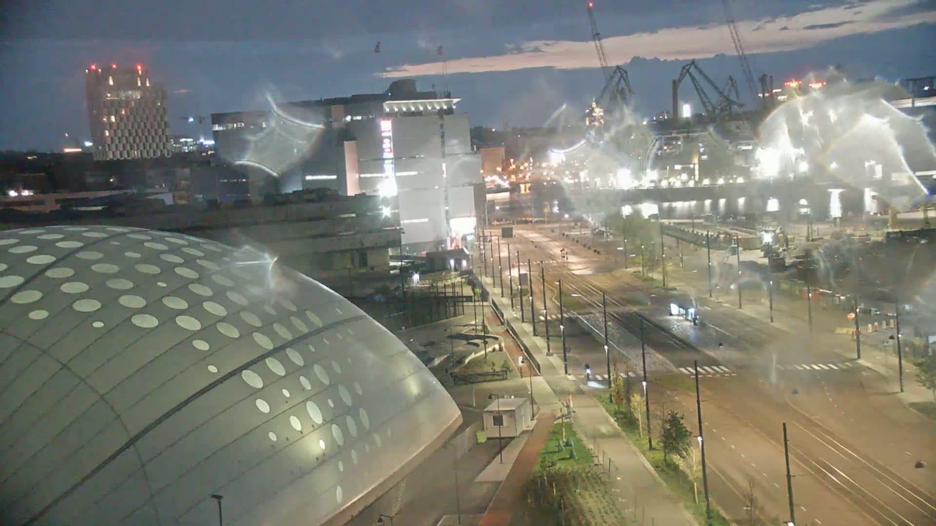 A nighttime cityscape with a large domed building in the foreground,  a roadway with tram tracks, and industrial areas and buildings in the background under a cloudy sky.