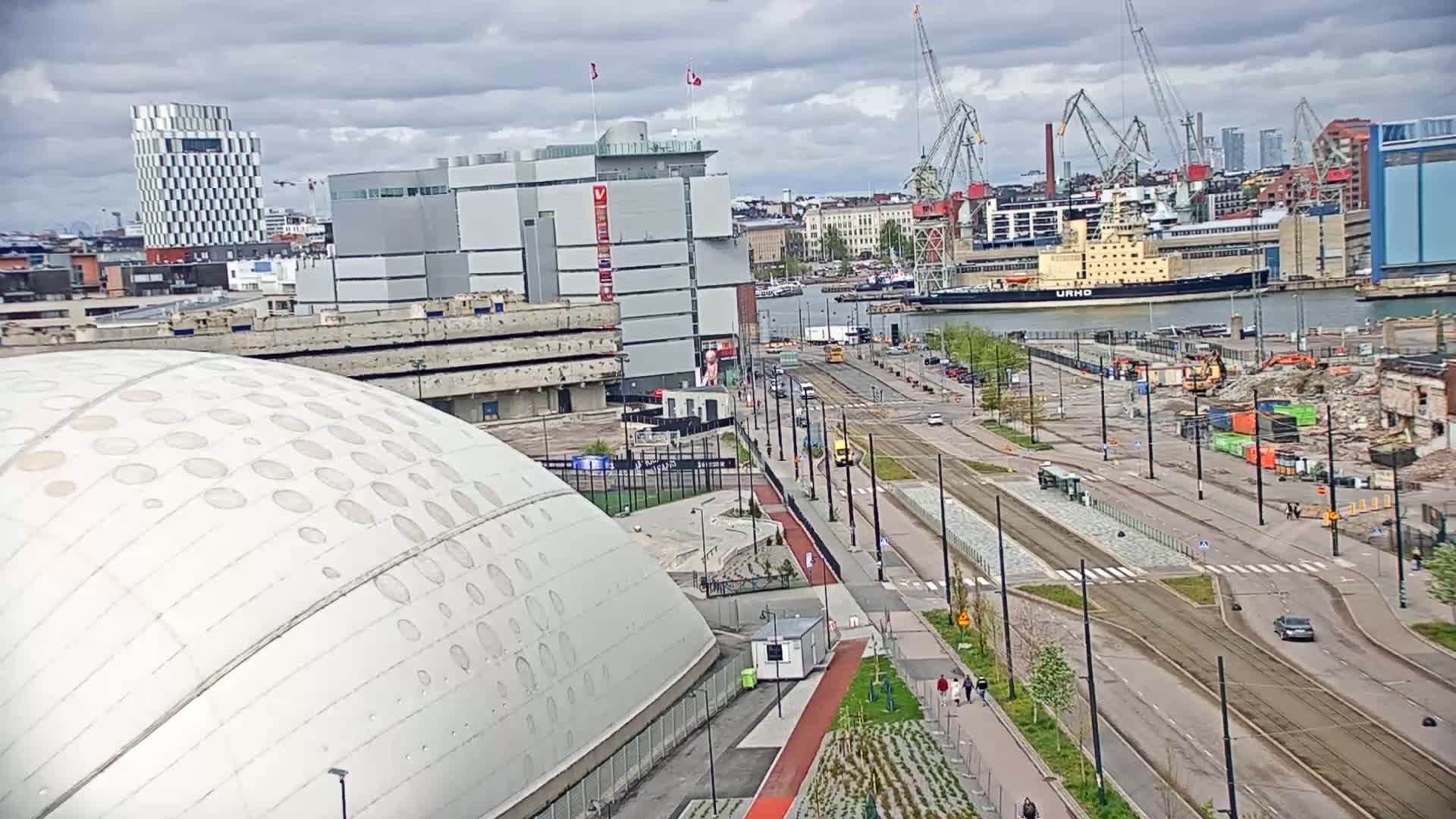 An elevated view shows a cityscape under cloudy skies, featuring a large domed building, a harbor with a ship, and a street with tram tracks undergoing construction.