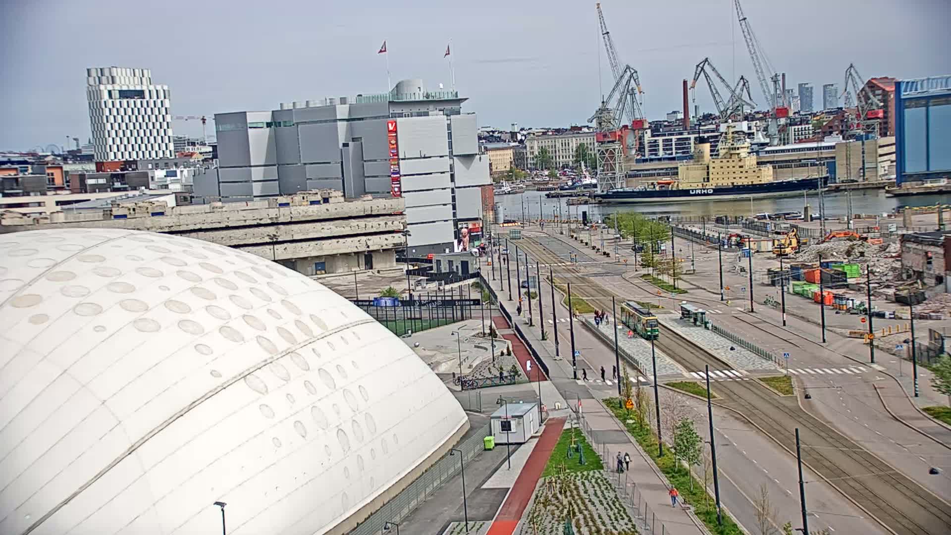 A large white dome-shaped building sits beside a harbor with several cranes and ships, under a mostly clear sky.