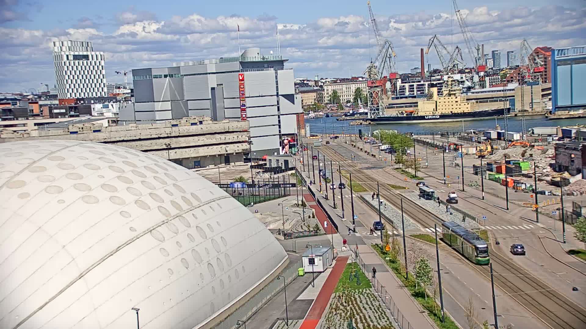 A partly cloudy day shows a cityscape with a large domed building in the foreground, a streetcar on a roadway, and a harbor with ships in the background.