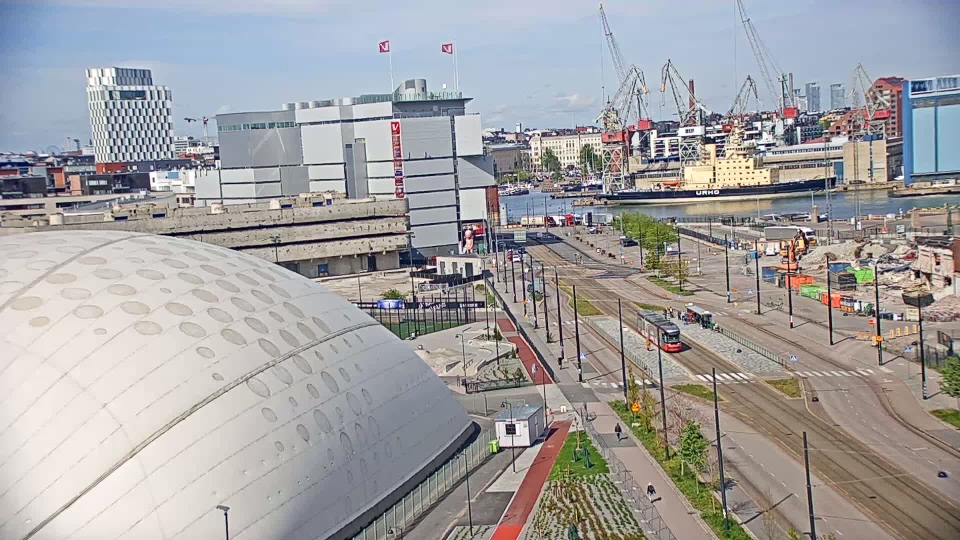 A large domed building with circular indentations is in the foreground, overlooking a city harbor with a large ship and tram tracks on a sunny day.