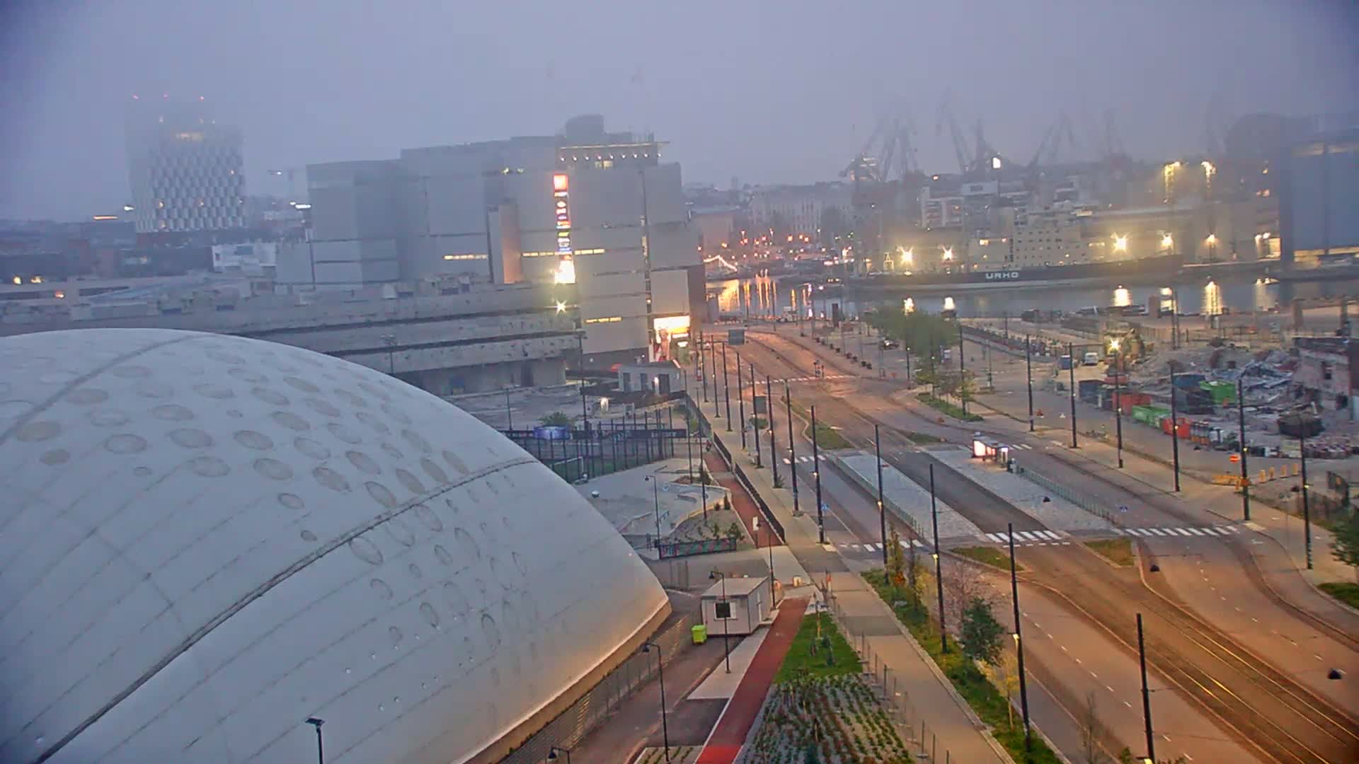 A large, domed building is situated near a wide, mostly empty road and tram tracks, with a hazy, dimly lit cityscape and a harbor in the background under a foggy sky.