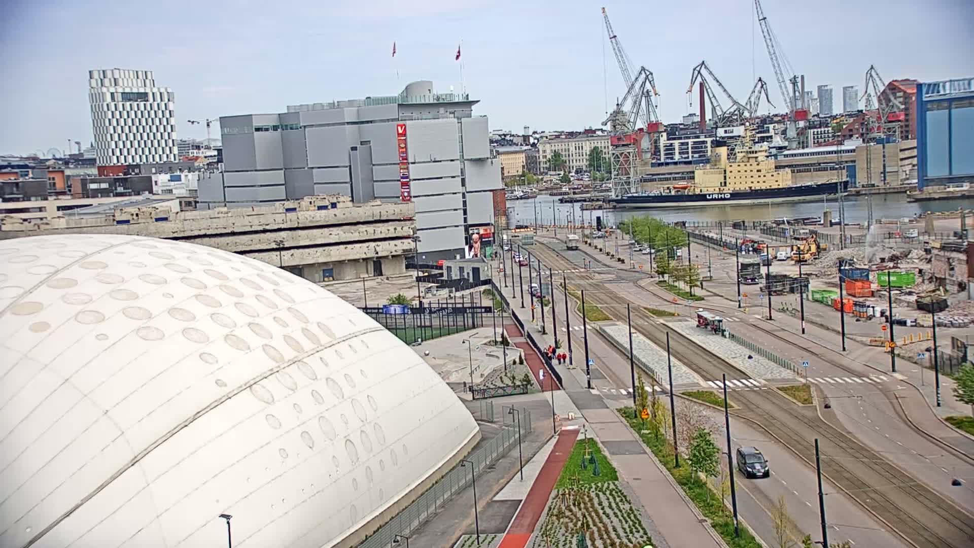 A large white dome-shaped building sits adjacent to a wide city street with tram lines, a harbor with a large ship, and ongoing demolition work under a partly cloudy sky.