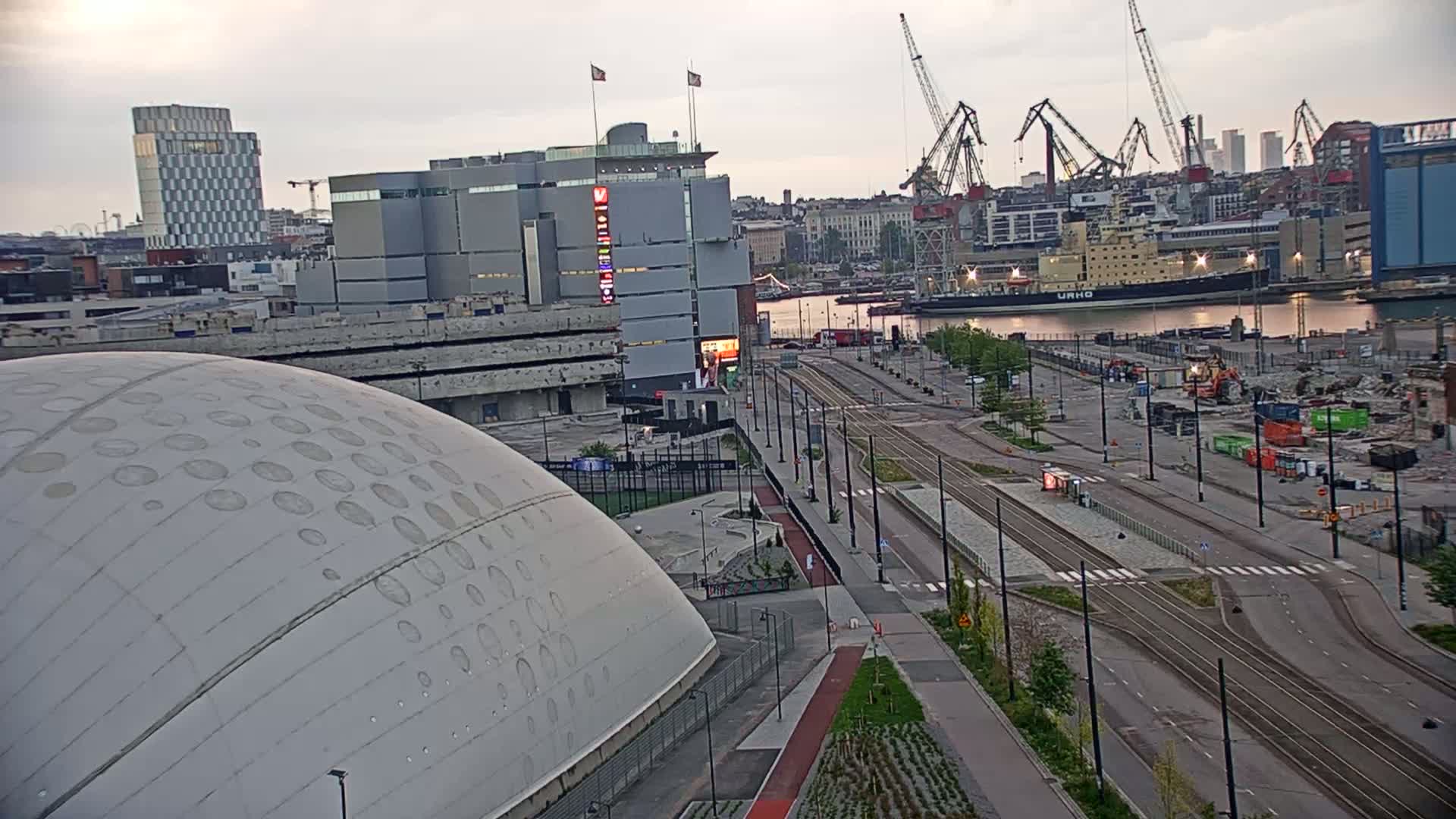 A large domed structure sits in the foreground of an overcast urban landscape, with tram lines running through a mostly empty plaza towards a harbor containing several large ships and cranes.
