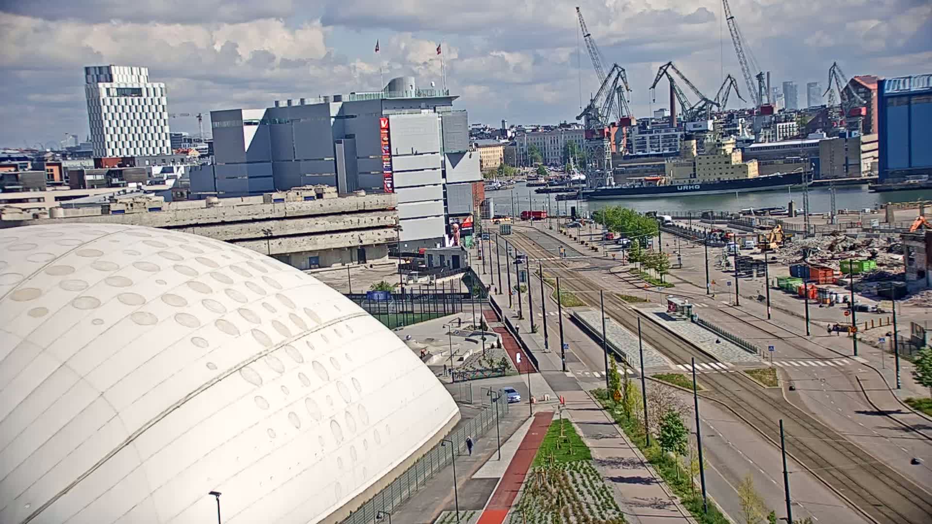 A partly cloudy day reveals a city harbor with large cranes, a cargo ship, and a large domed building next to a tram line and construction activity.