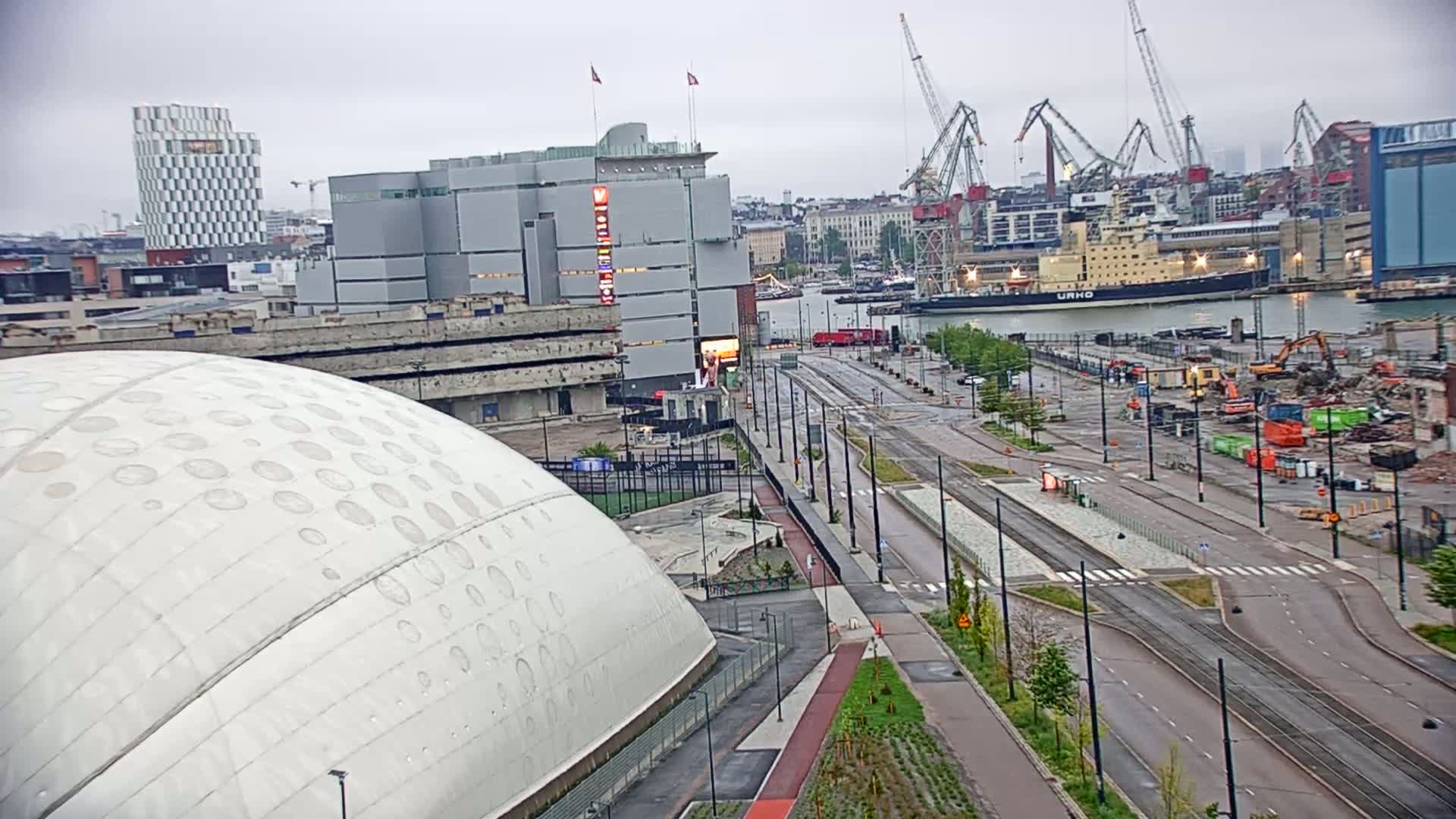 A large domed building sits beside a wide roadway with tram lines running through a city harbor area under overcast skies.