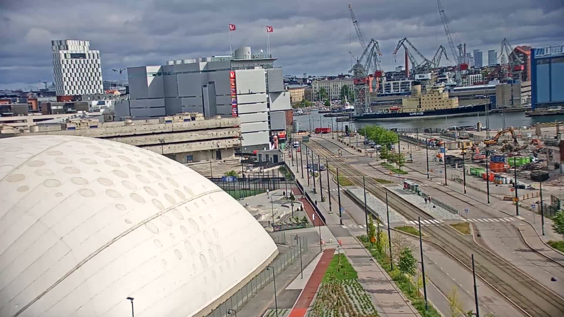 A large white domed building is situated next to a wide city street with tram lines, under a cloudy sky, overlooking a harbor with ships and cranes.