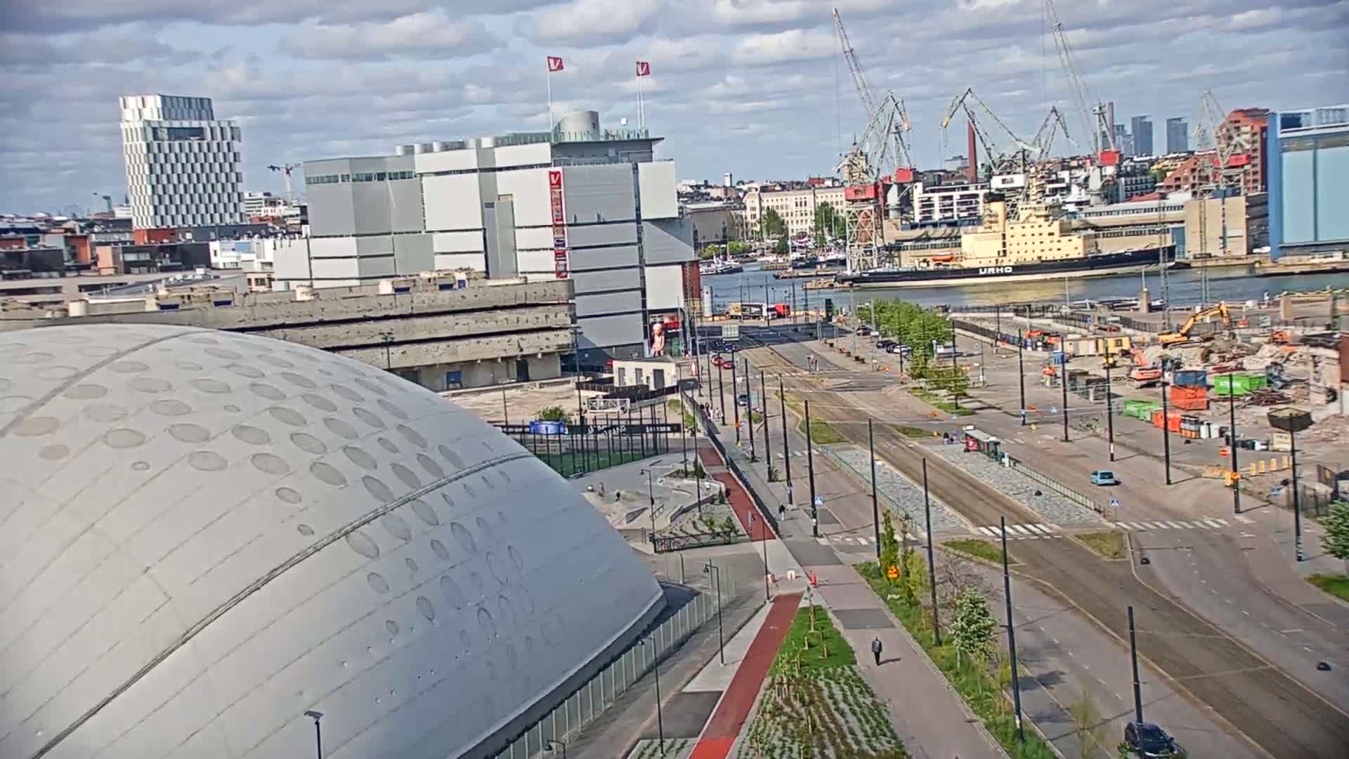 A partly cloudy day reveals an urban waterfront scene with a large domed building in the foreground, a harbor with industrial cranes and ships, and a city skyline in the background; demolition is visible in the midground.
