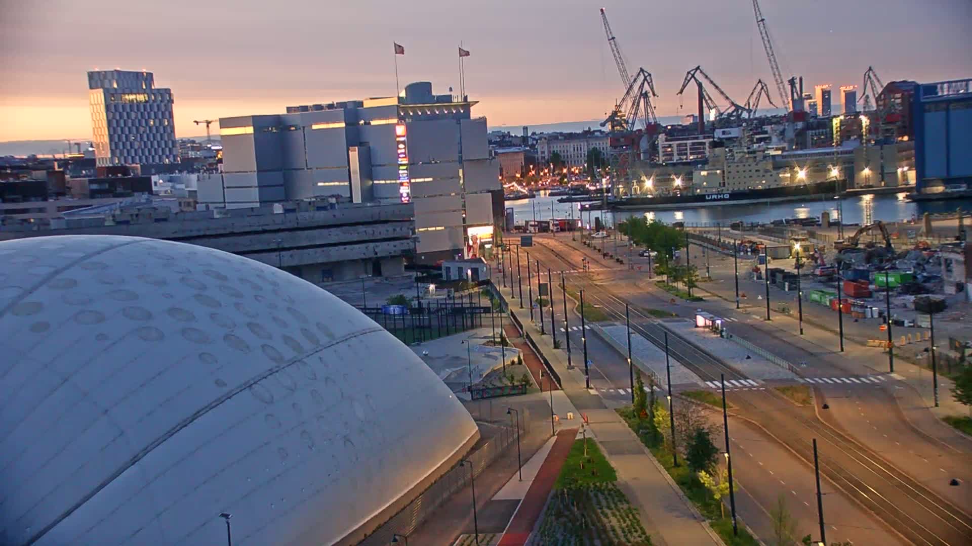 A city harbor at dusk, featuring a large domed building, modern architecture, a busy port with cranes and ships, and empty streets with streetcars.
