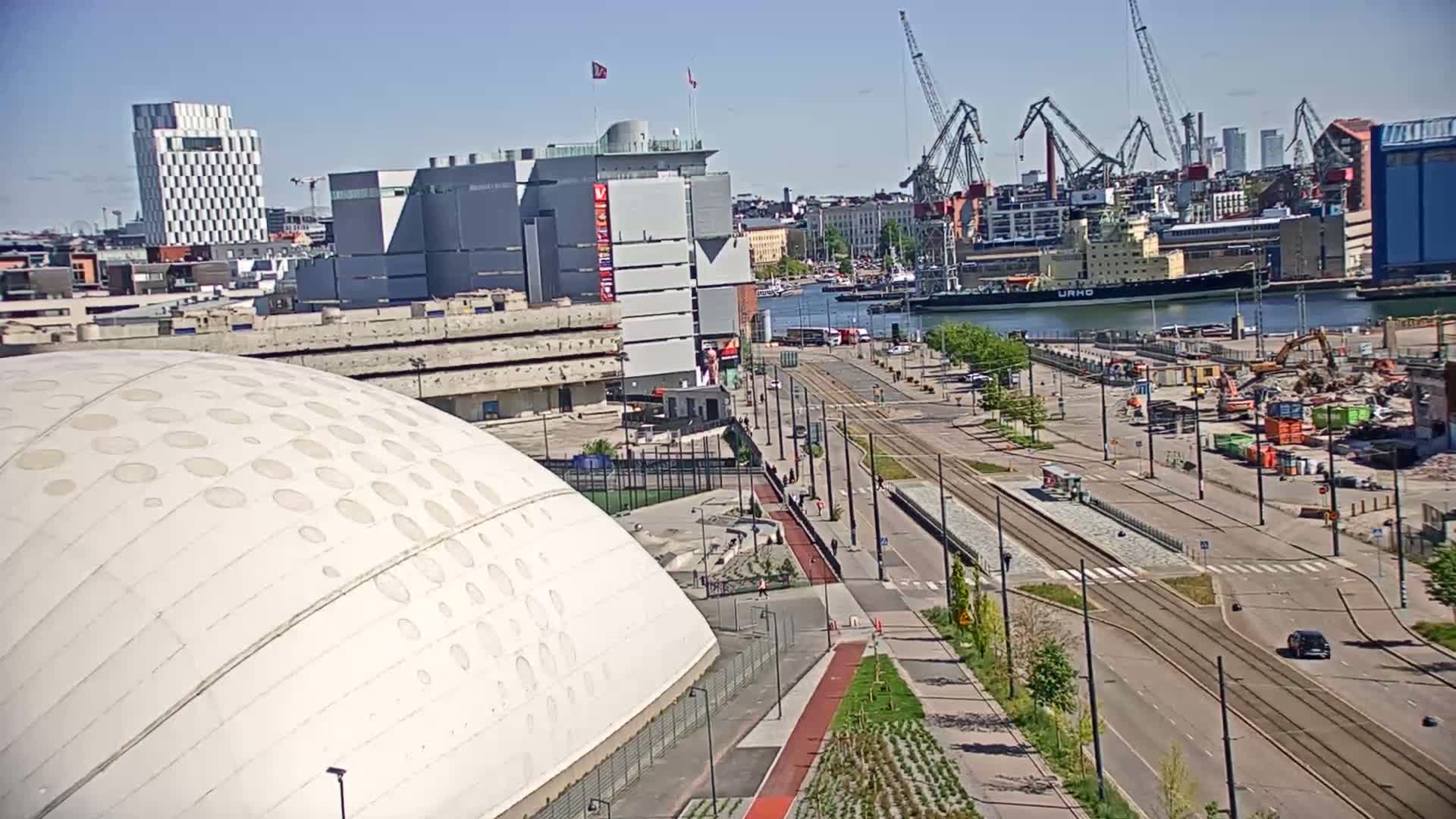 A large white domed building is situated near a waterfront with construction, tram tracks, and several cranes visible on a sunny day.