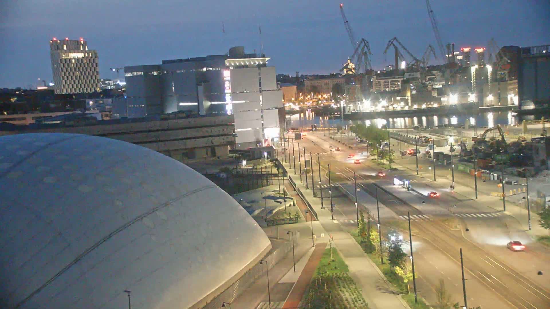 A nighttime, high-angle view shows a large, dome-shaped building next to a multi-lane roadway with sparse traffic, and a harbor with industrial cranes and buildings in the background under a twilight sky.