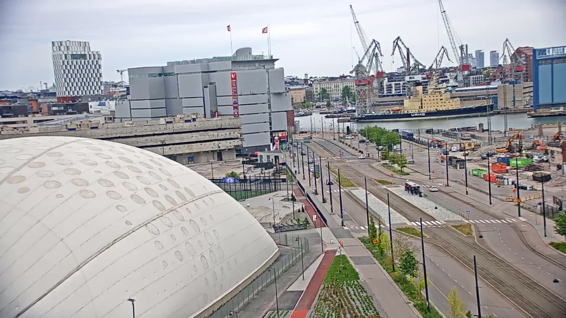 A high-angle view shows a city harbor with docked ships under an overcast sky, featuring a large domed building and a wide road with tram tracks leading towards the water.