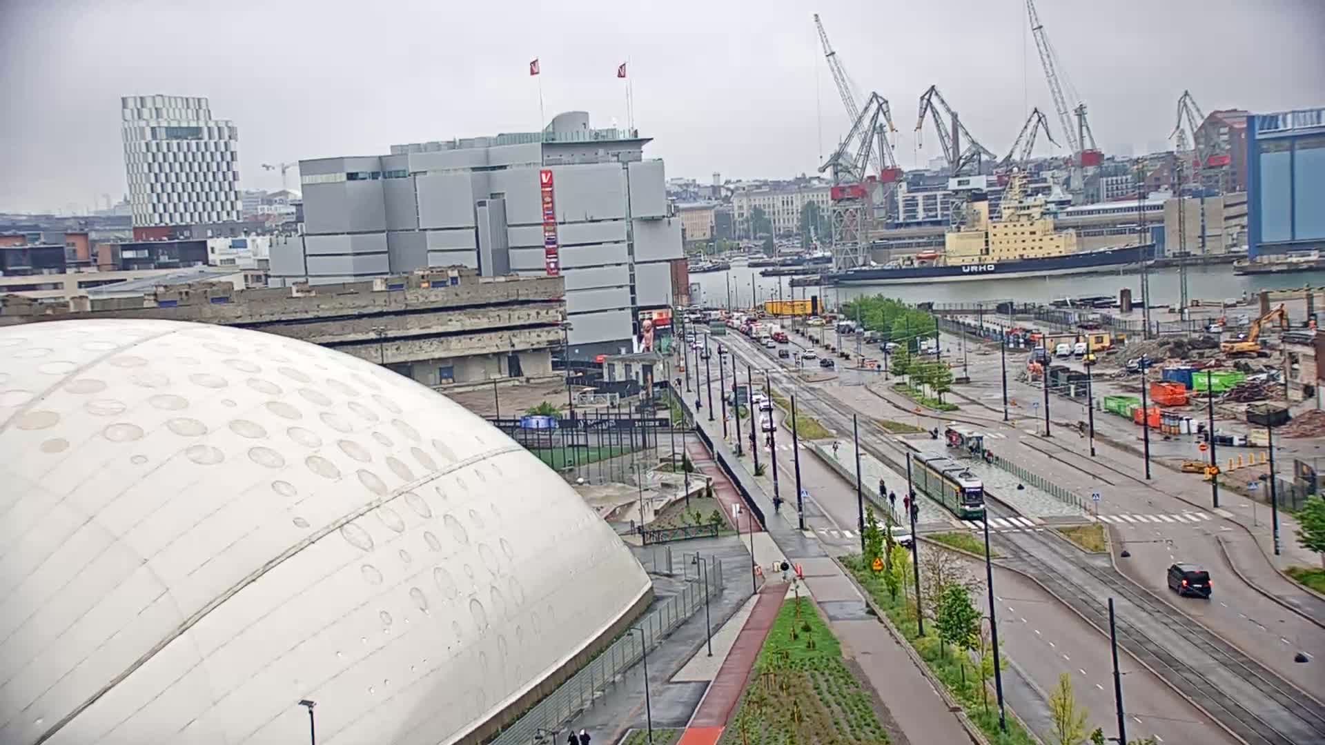 A large, domed building sits adjacent to a roadway with a tram, harbor, and construction site under an overcast sky.