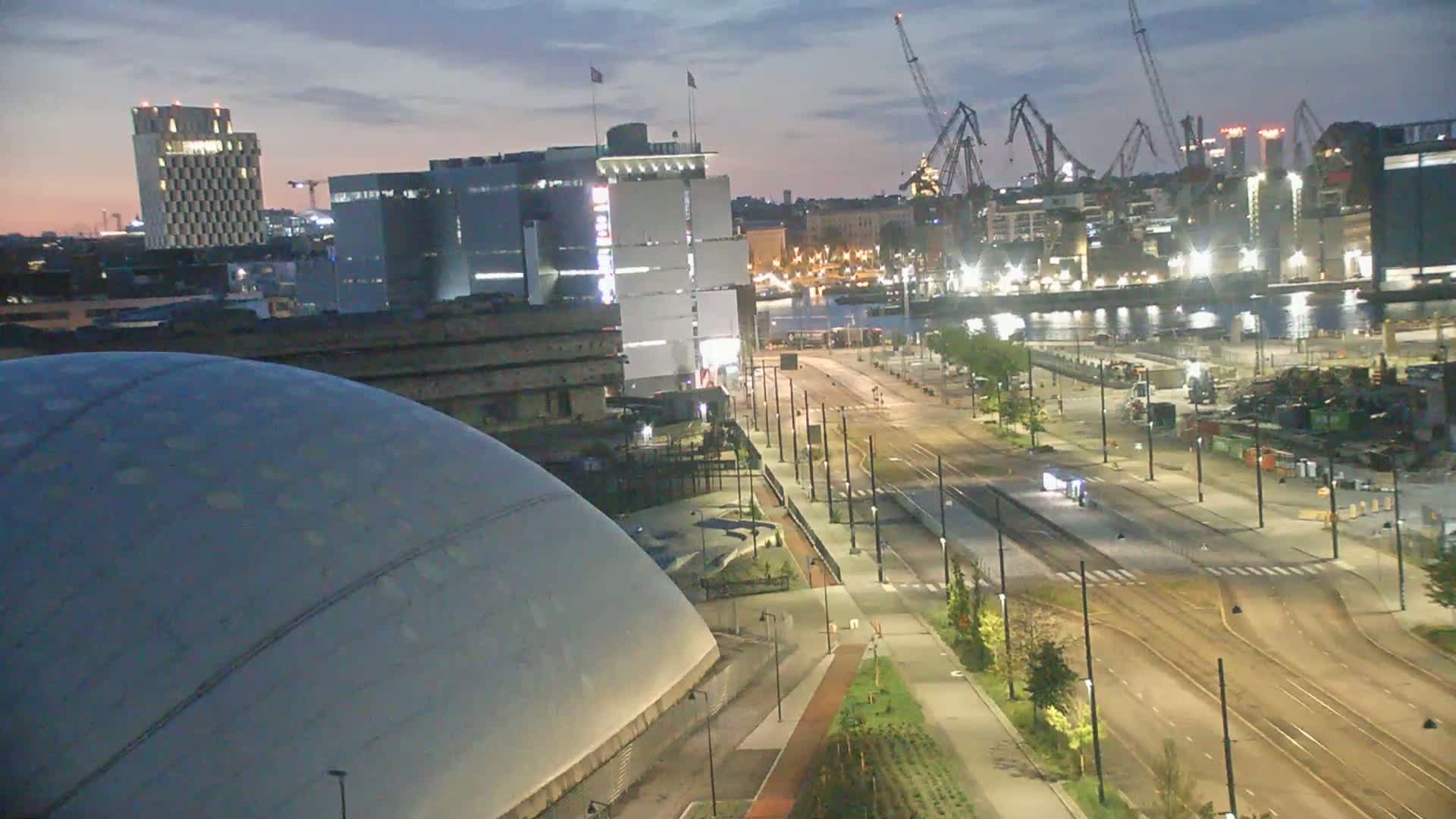 A nighttime, high-angle view of a city harbor with a large dome-shaped building in the foreground,  empty streets and tram tracks, and cranes in the background under a twilight sky.