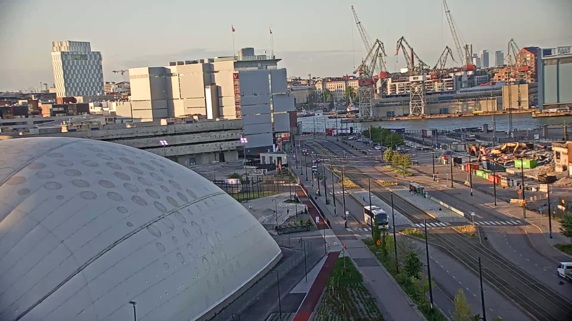 A large, domed building is situated near a waterfront city with several cranes visible in the distance under a partly sunny sky.