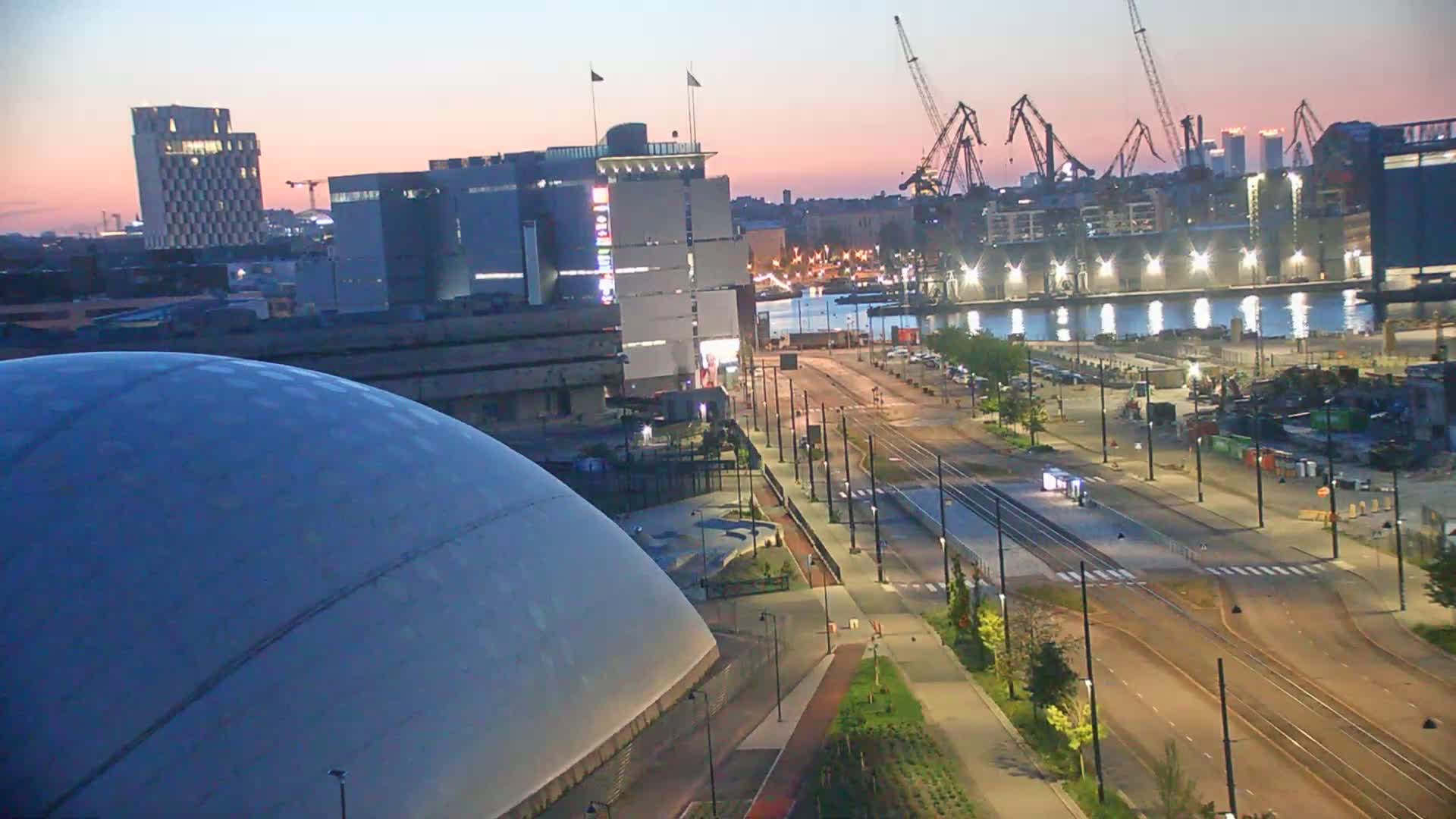 A large, domed building is situated near a waterfront area with urban infrastructure, including tram lines and industrial cranes, at dusk under a clear sky.