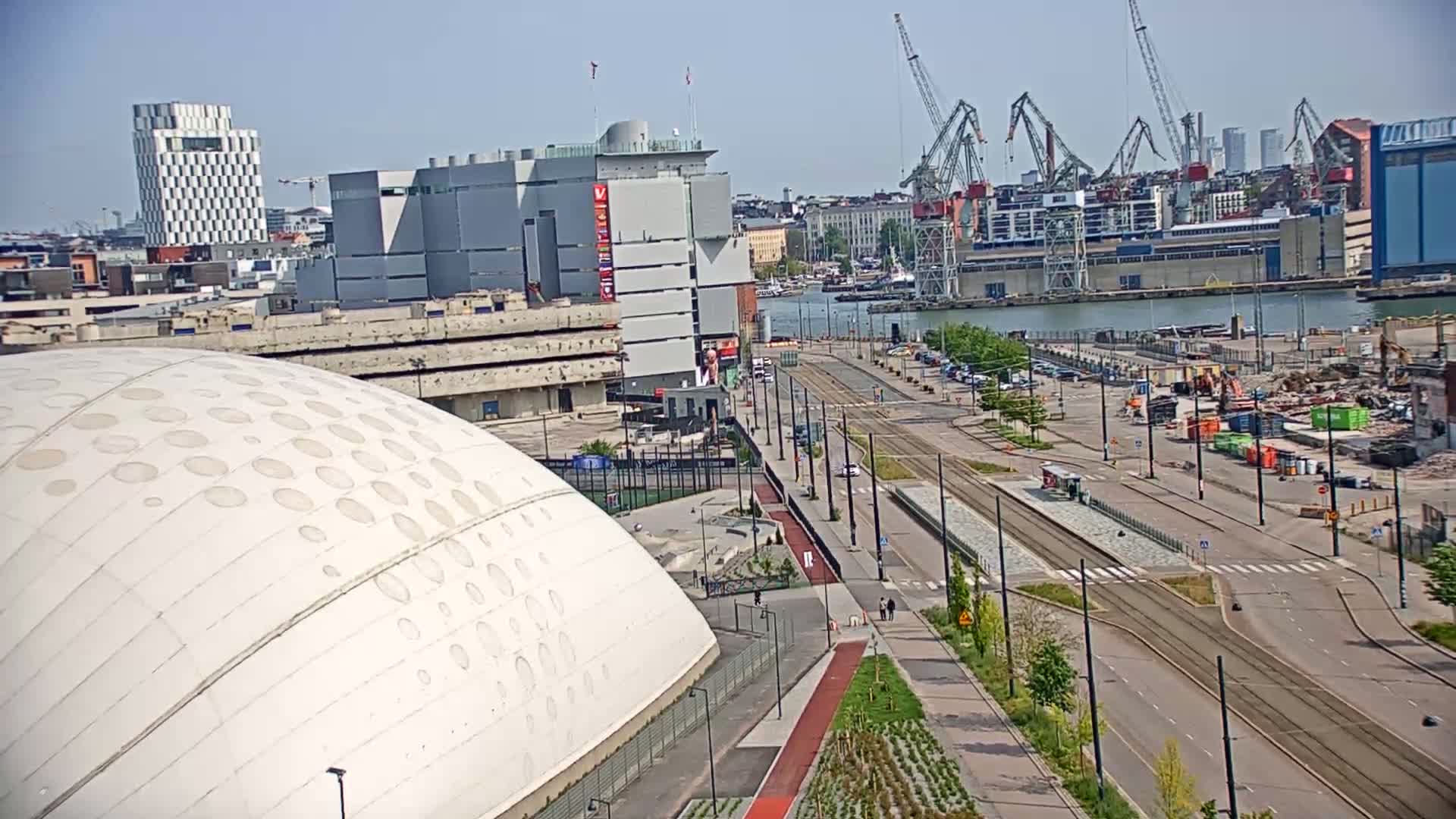 A large, white domed building is visible in the foreground, next to a streetcar line and a waterway with industrial buildings and cranes in the background, under a clear, sunny sky.