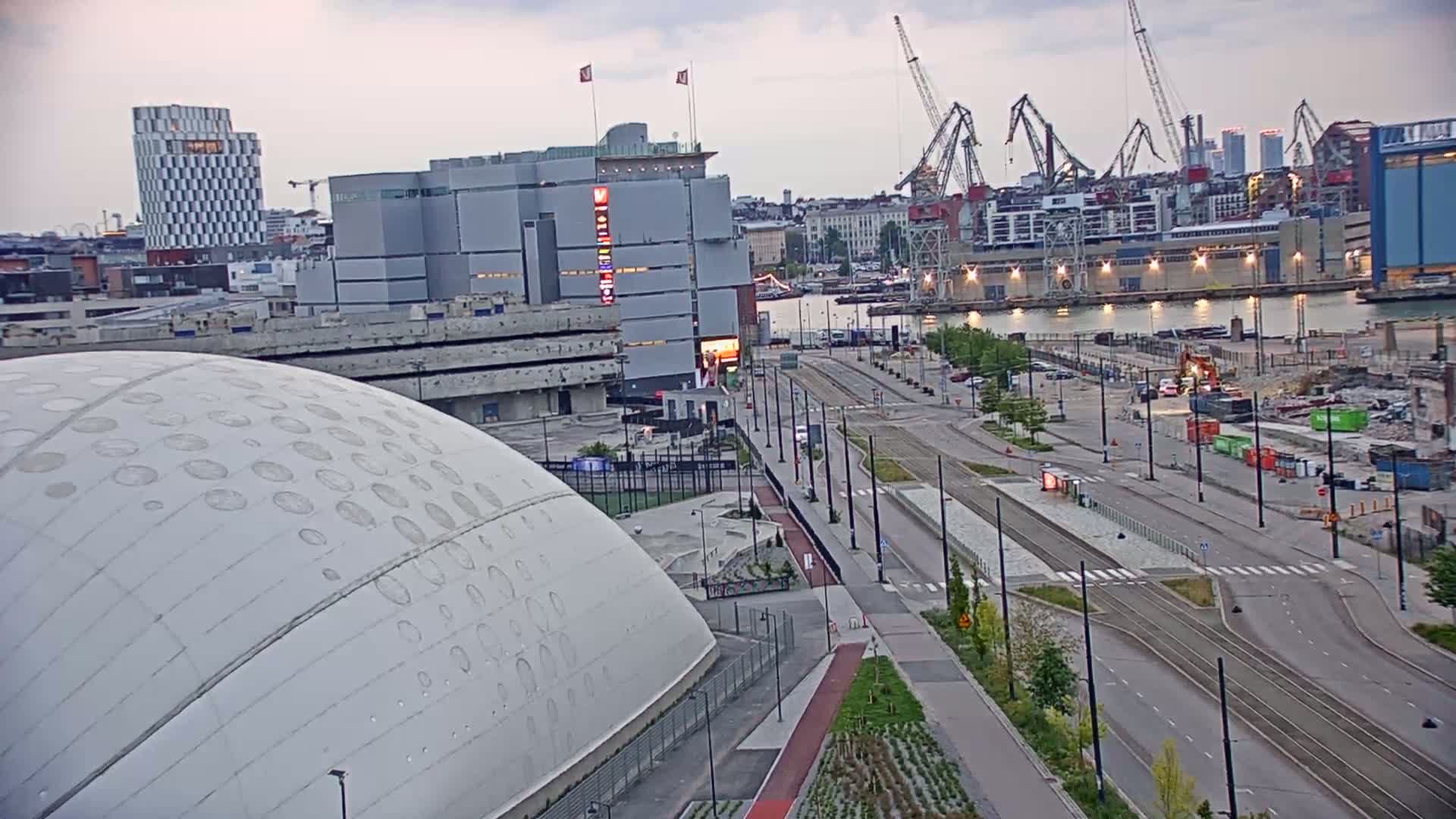A large, domed building sits beside a wide road and tram tracks, overlooking a harbor with cranes and several buildings under an overcast sky.