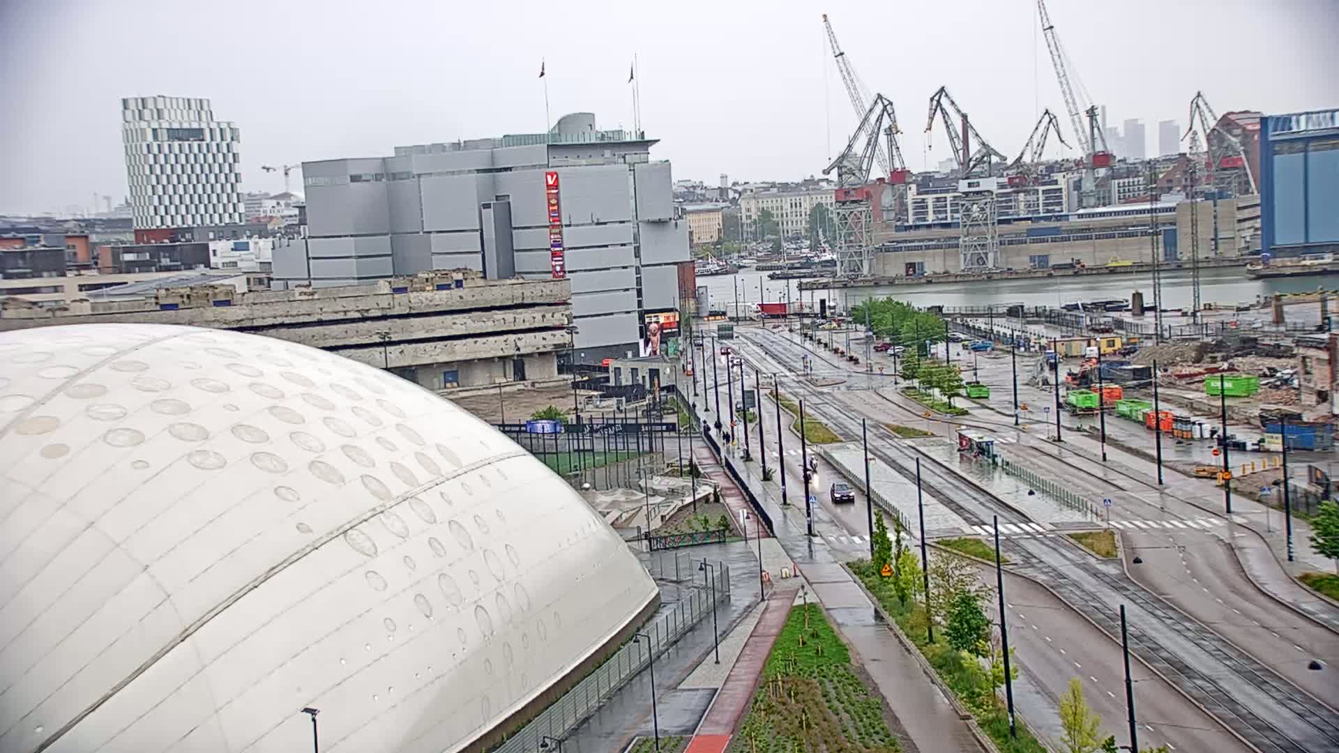 A large domed building sits beside a wet city street with tram tracks and construction visible in the background under a cloudy sky.