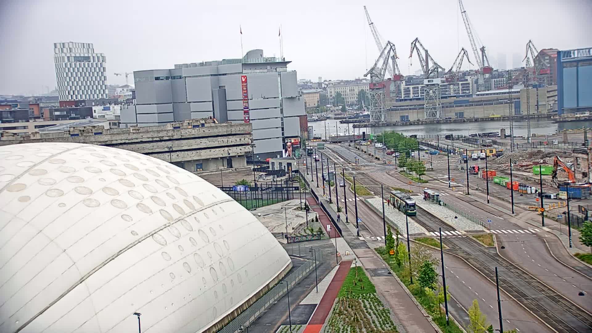 A white domed building is seen in the foreground with a city landscape and a body of water beyond, under an overcast sky.