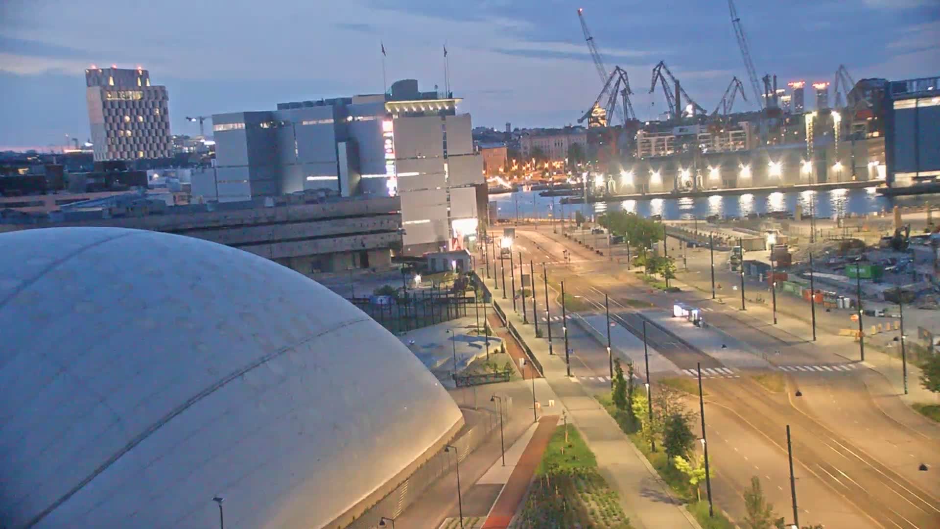 A large, domed building sits near a waterfront at twilight, with a wide, mostly empty road and landscaped median separating it from a harbor containing several cranes and industrial buildings.