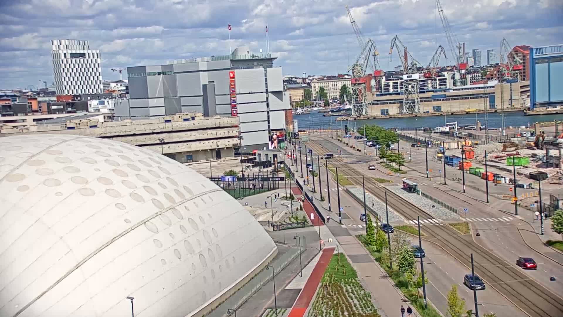 A large, domed building with a patterned surface is situated near a waterfront area with tram tracks and construction activity under a partly cloudy sky.