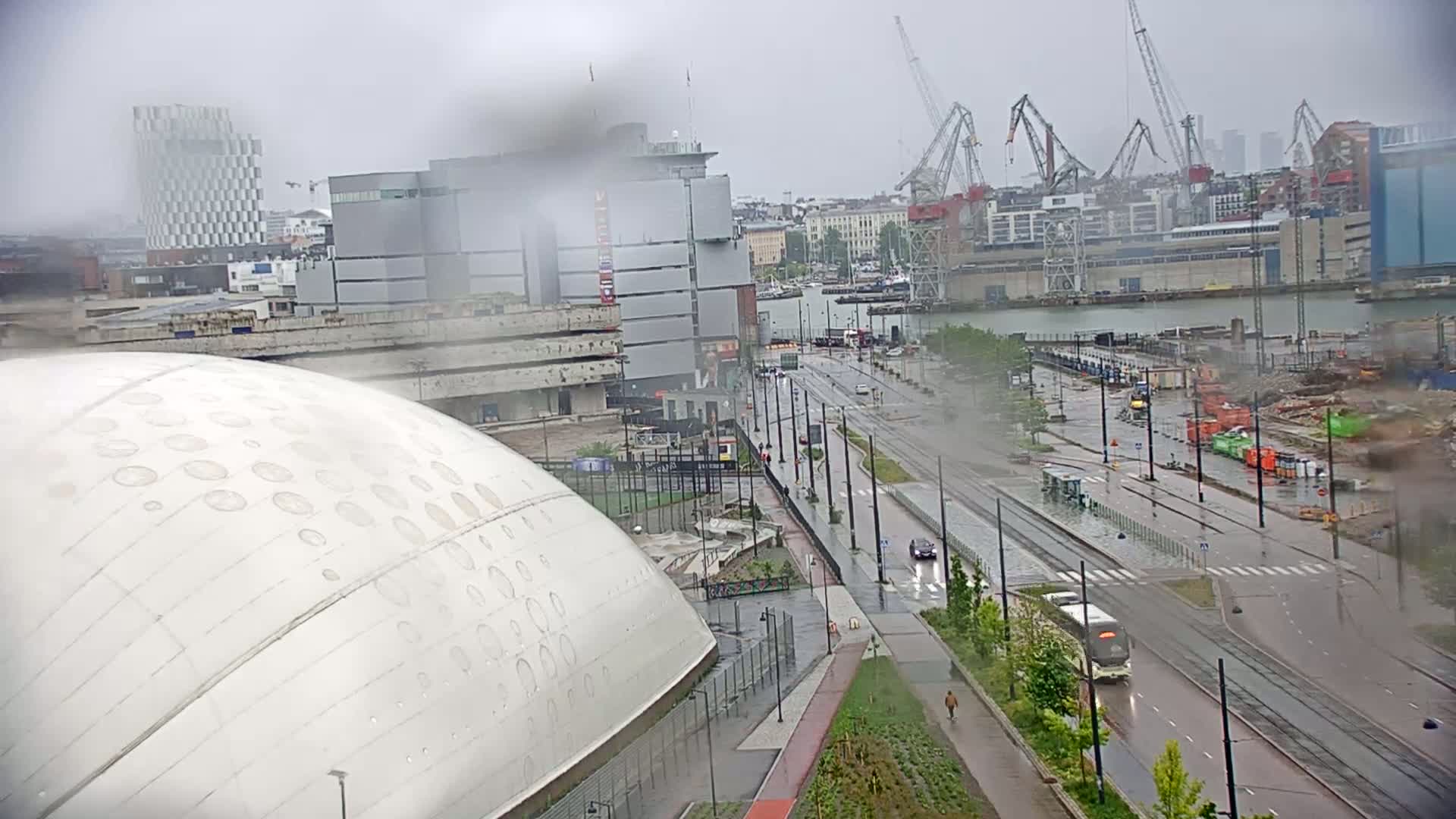 A wet city street scene, viewed from above, shows a large domed building, a tram line, and a harbor with cranes under a rainy sky.
