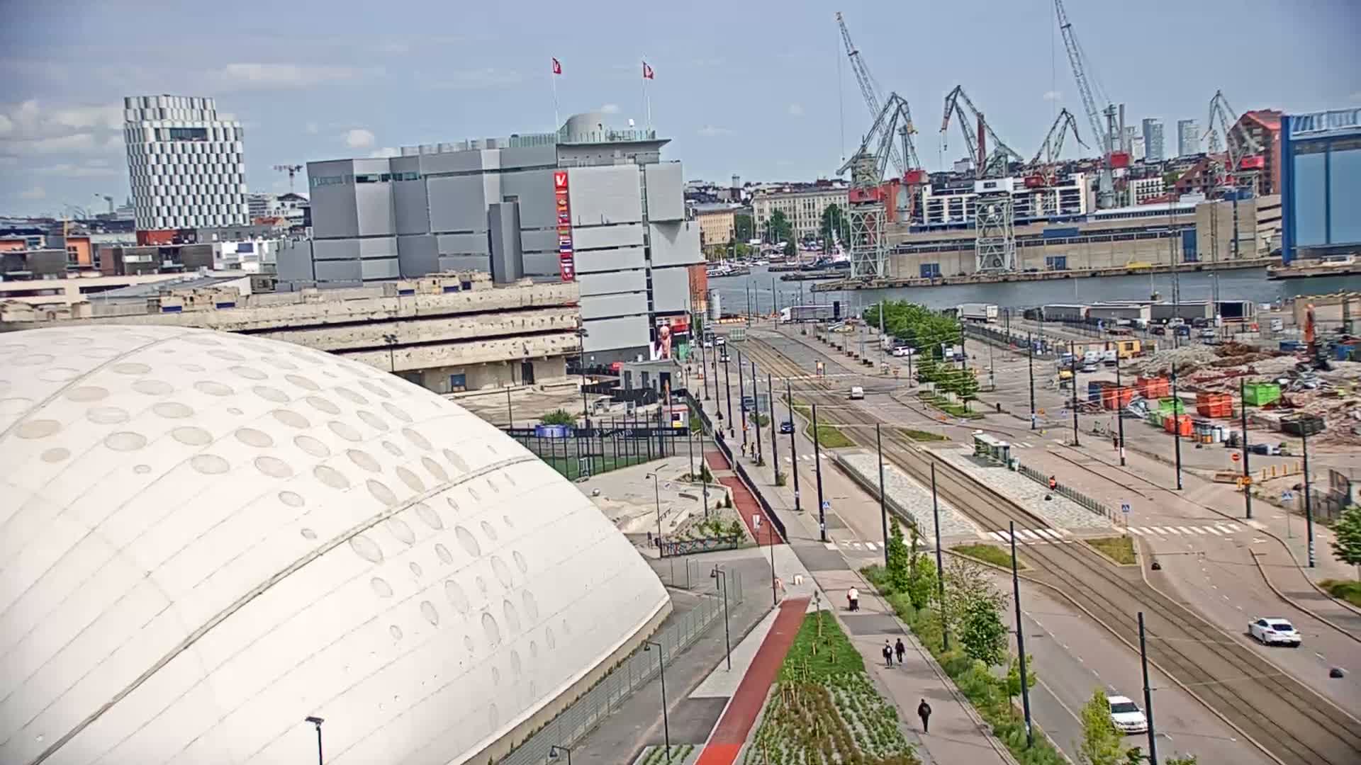 A large domed building is situated next to a harbor with multiple cranes and urban development, under a mostly sunny sky.