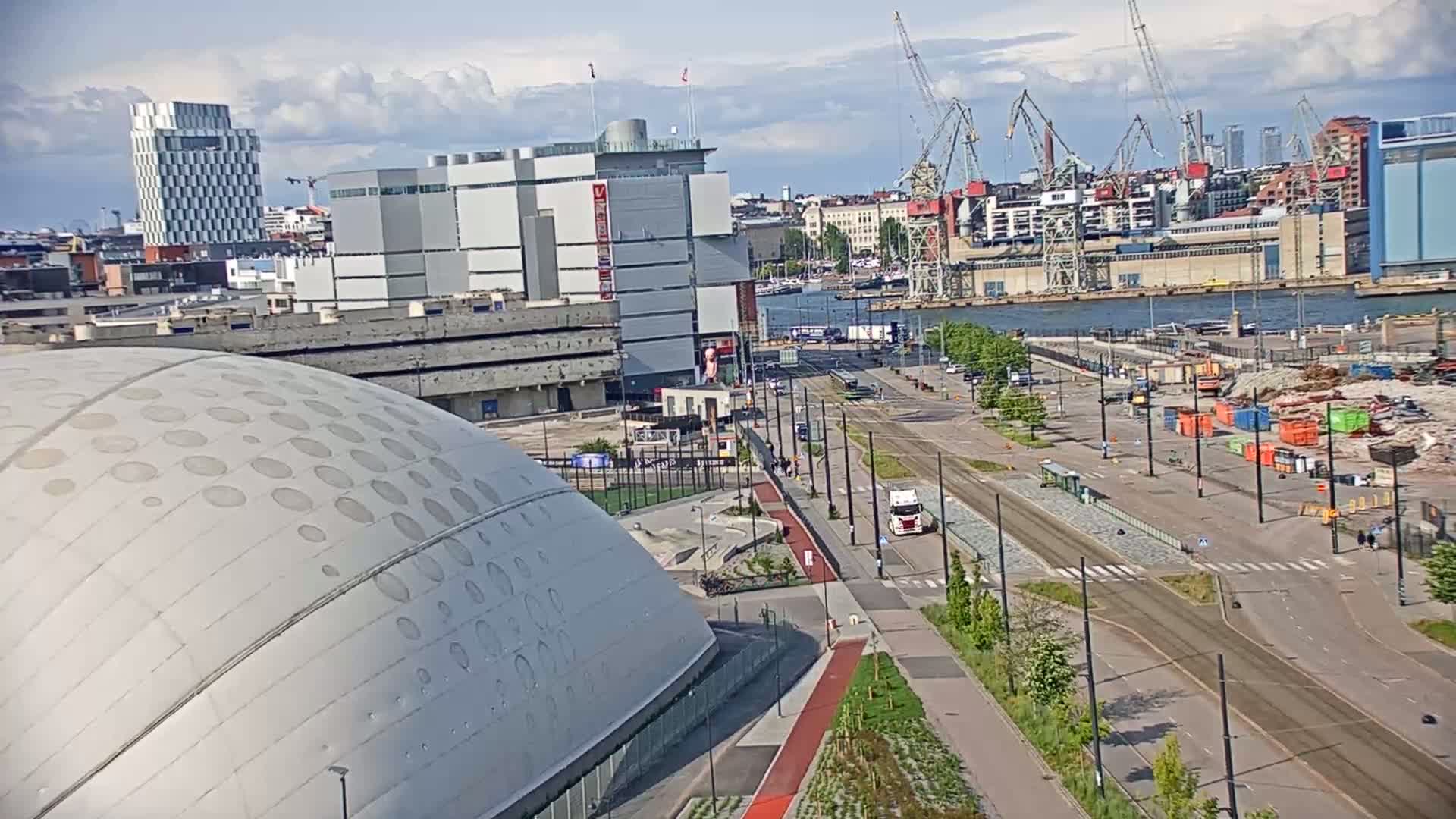 A large domed building is seen near a waterfront area with a harbor, cranes, and several buildings under a partly cloudy sky.