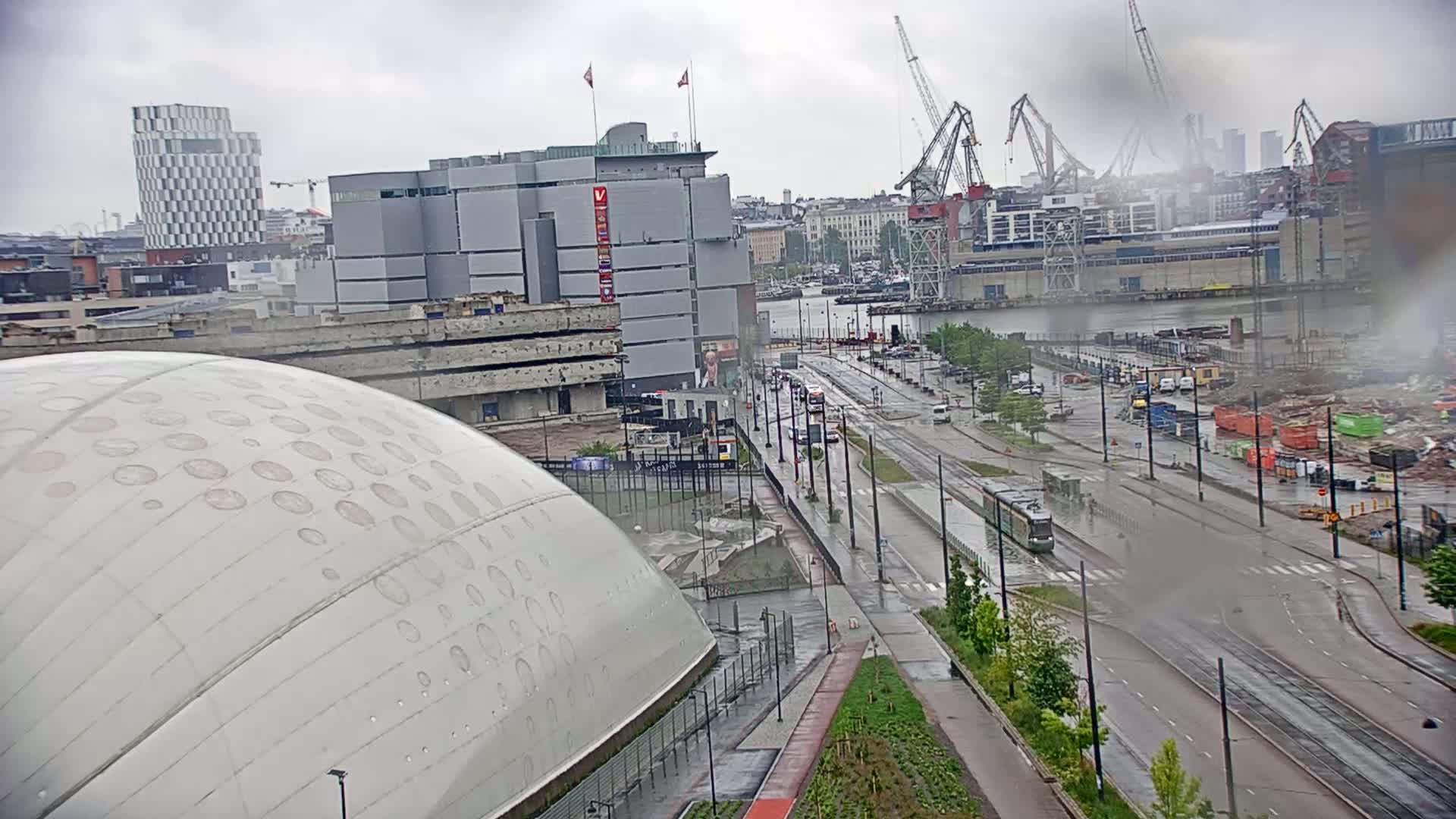 A rain-soaked cityscape features a large domed building, modern architecture, a tram on a street, and construction activity near a waterfront.