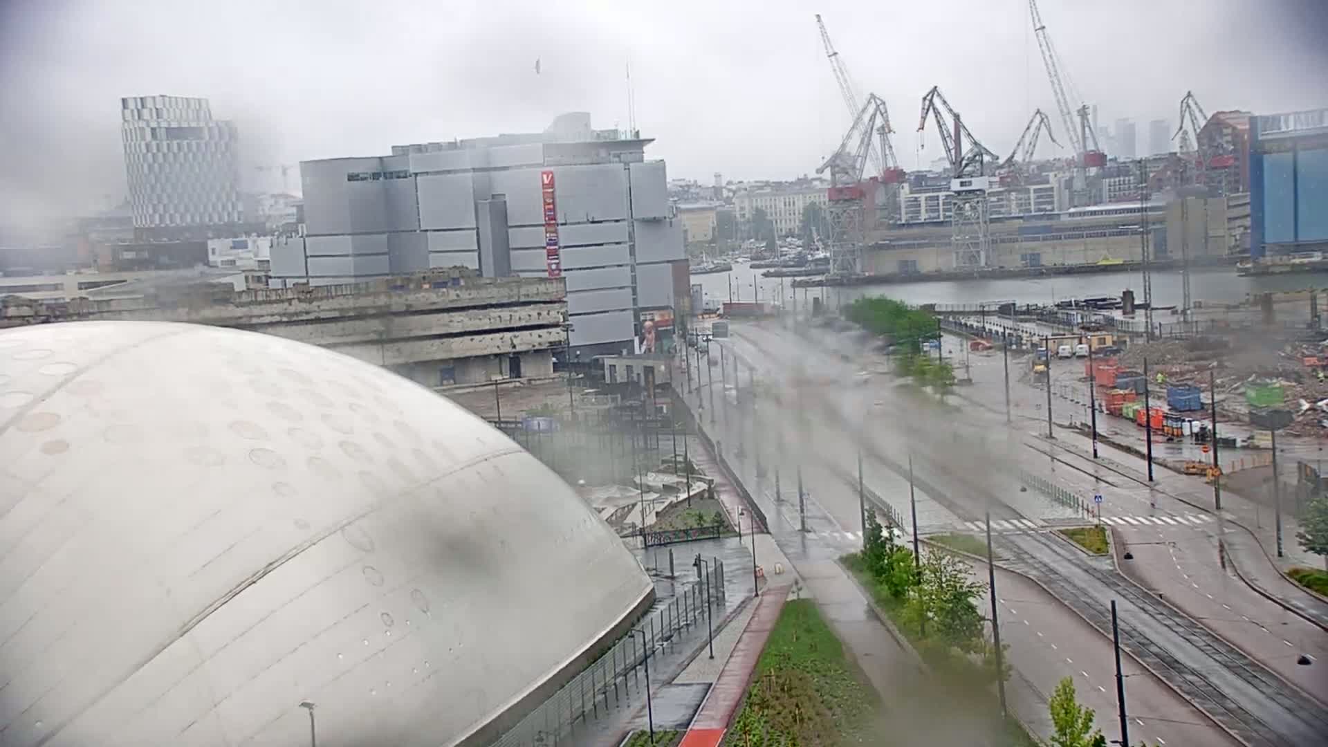 A rain-swept city scene shows a large domed building next to a street with tram tracks, passing by a construction site and harbor with cranes under an overcast sky.