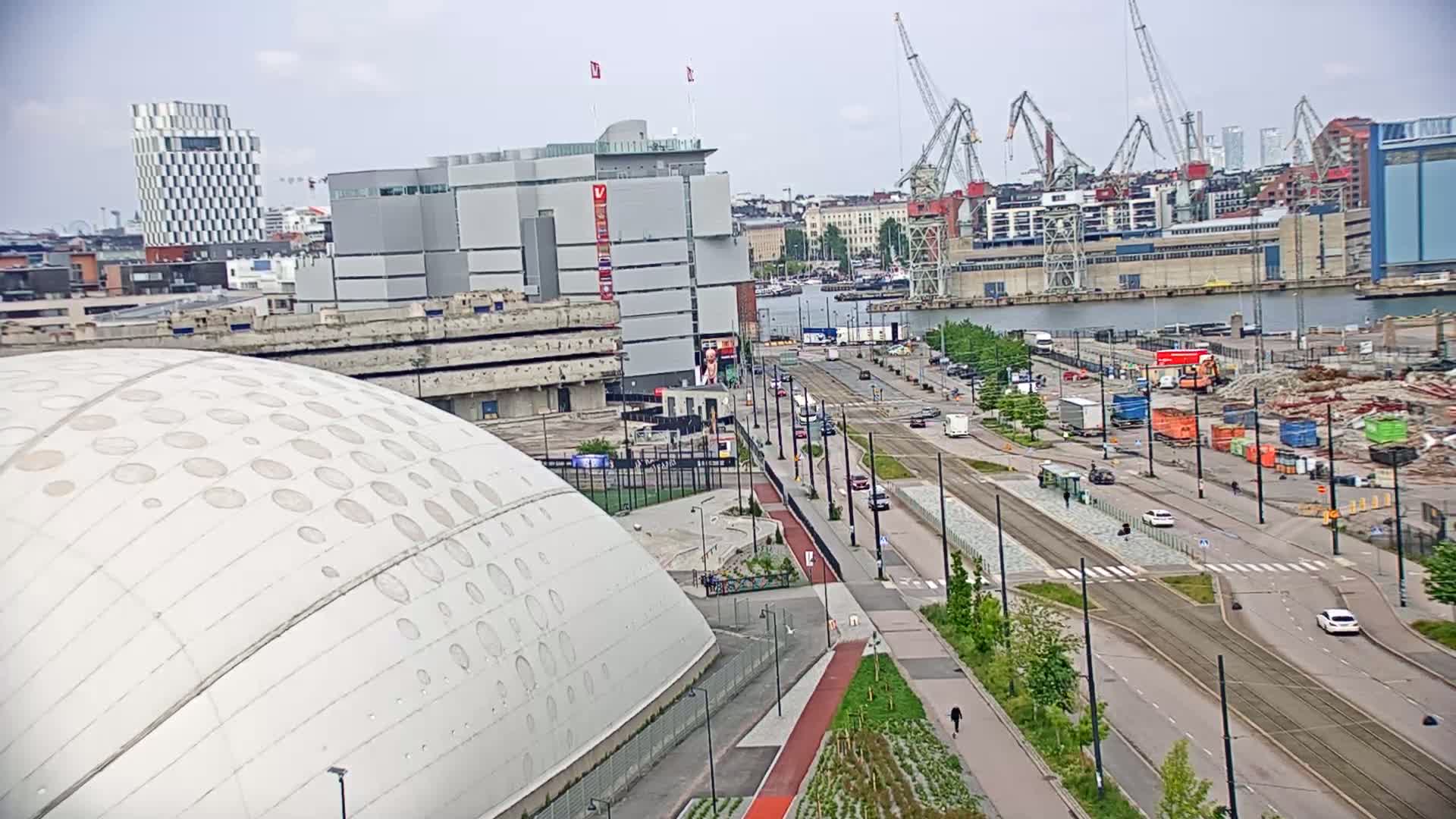 A large domed building is situated near a waterfront area with several cranes, buildings, and vehicles on a partly cloudy day.