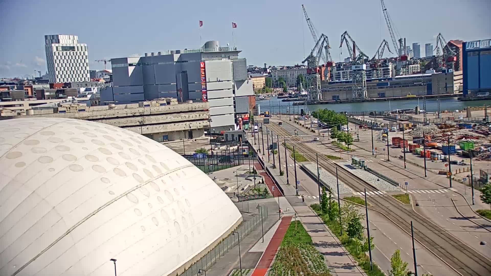 A large, domed building with circular indentations is situated near a waterfront area on a sunny day, with tram lines and construction visible in the background.