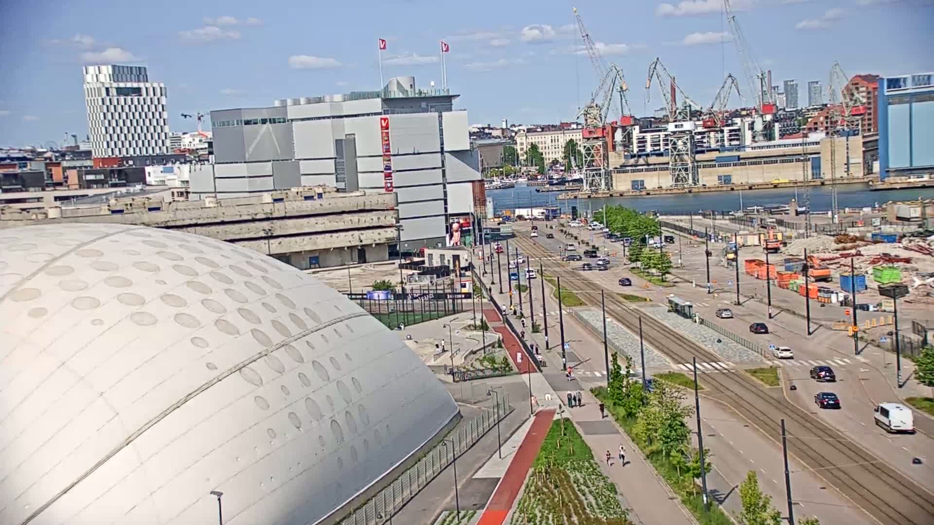 A large domed building sits near a waterfront city on a sunny day, with a tram line and several cars visible on a nearby street.