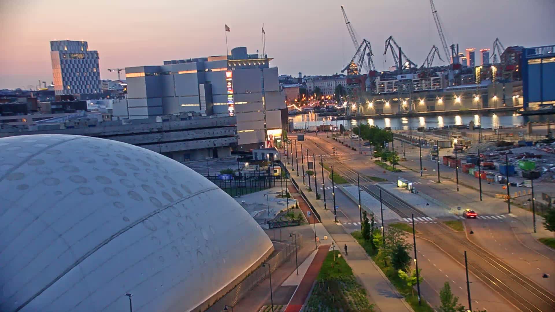 A large, domed building sits next to a wide, empty roadway at dusk, with a body of water, industrial cranes, and several buildings in the background.