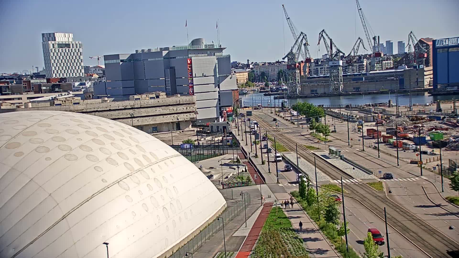 A large, domed building sits next to a wide, empty roadway at dusk, with a body of water, industrial cranes, and several buildings in the background.