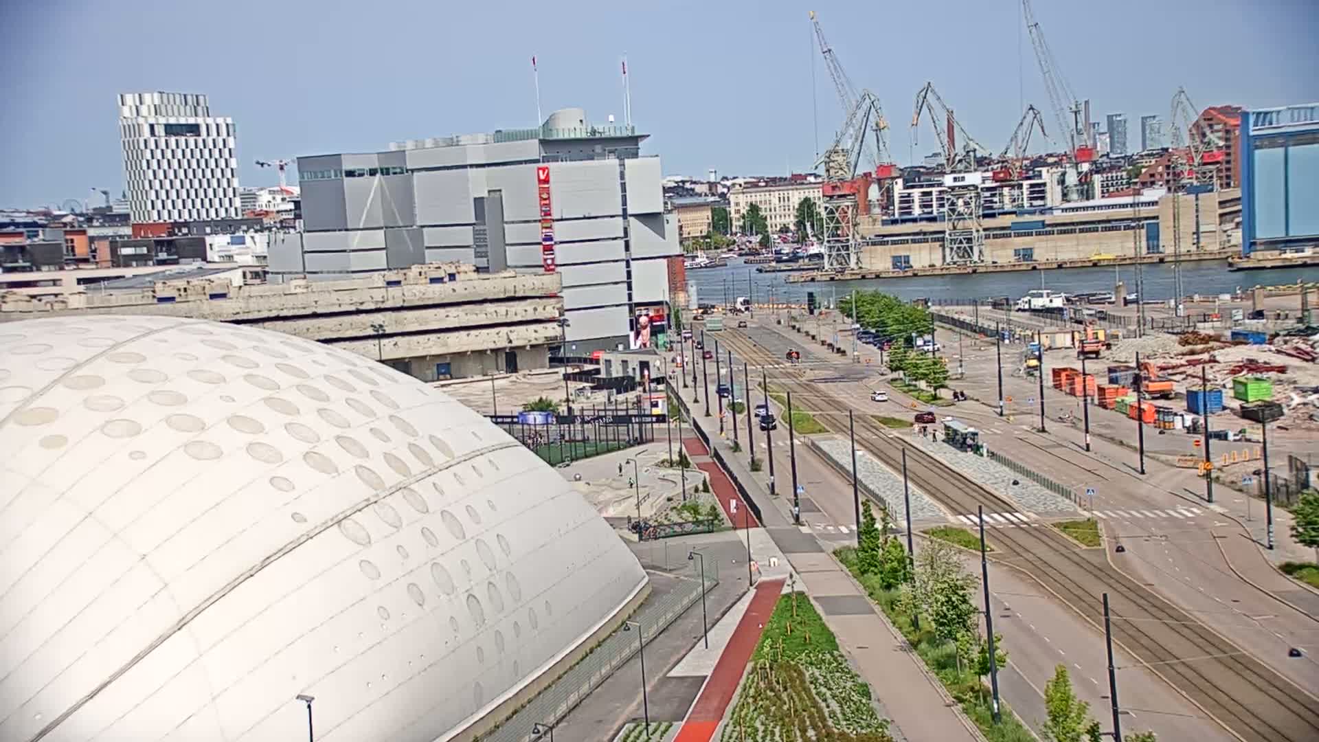 A large, domed building with circular indentations sits beside a city street with tram tracks running through it, under a clear, sunny sky, and across from a harbor with cranes and construction.