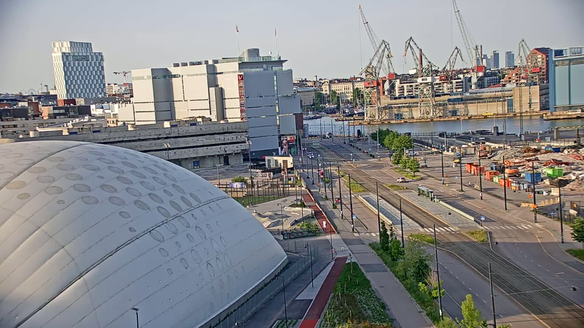 A large domed building sits near a harbor in a city on a clear, sunny day, with tram lines running through a street in front of it.