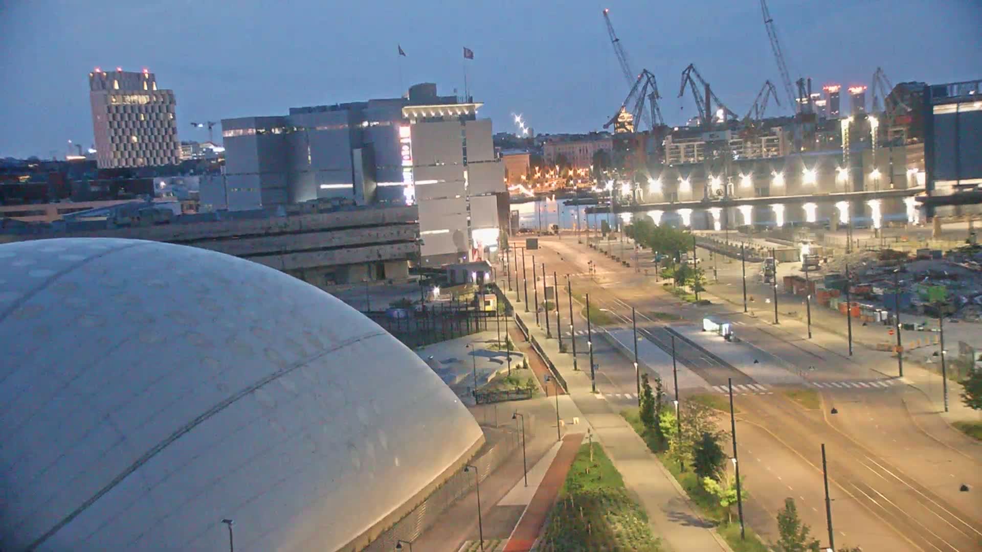 A large domed building sits near a harbor in a city on a clear, sunny day, with tram lines running through a street in front of it.