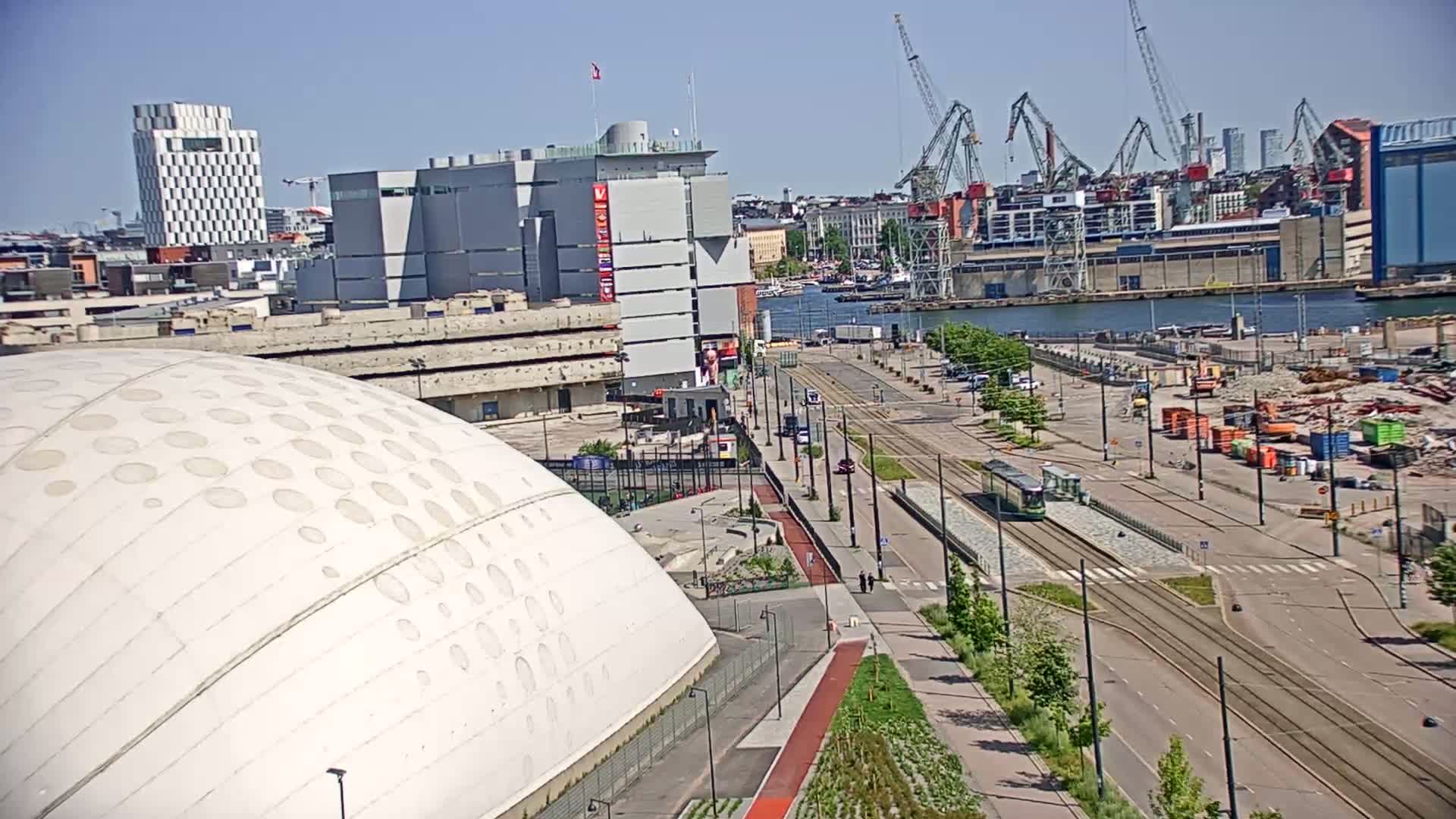 A large white dome-shaped structure sits beside a harbor on a sunny day, with a tram running along a roadway in the foreground and cranes visible in the distance.