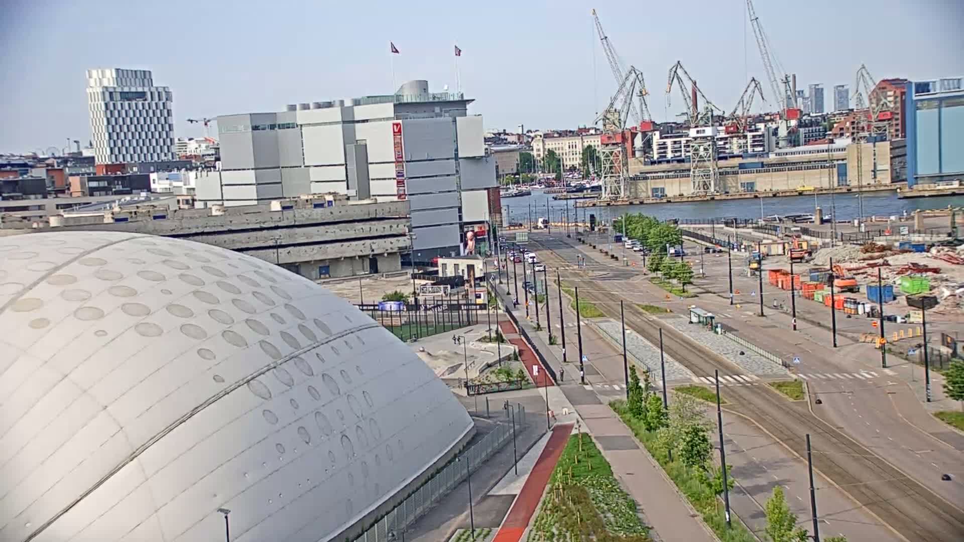 A large domed building sits next to a wide roadway with sparse traffic on a sunny day, overlooking a harbor with several cranes and a demolition site.
